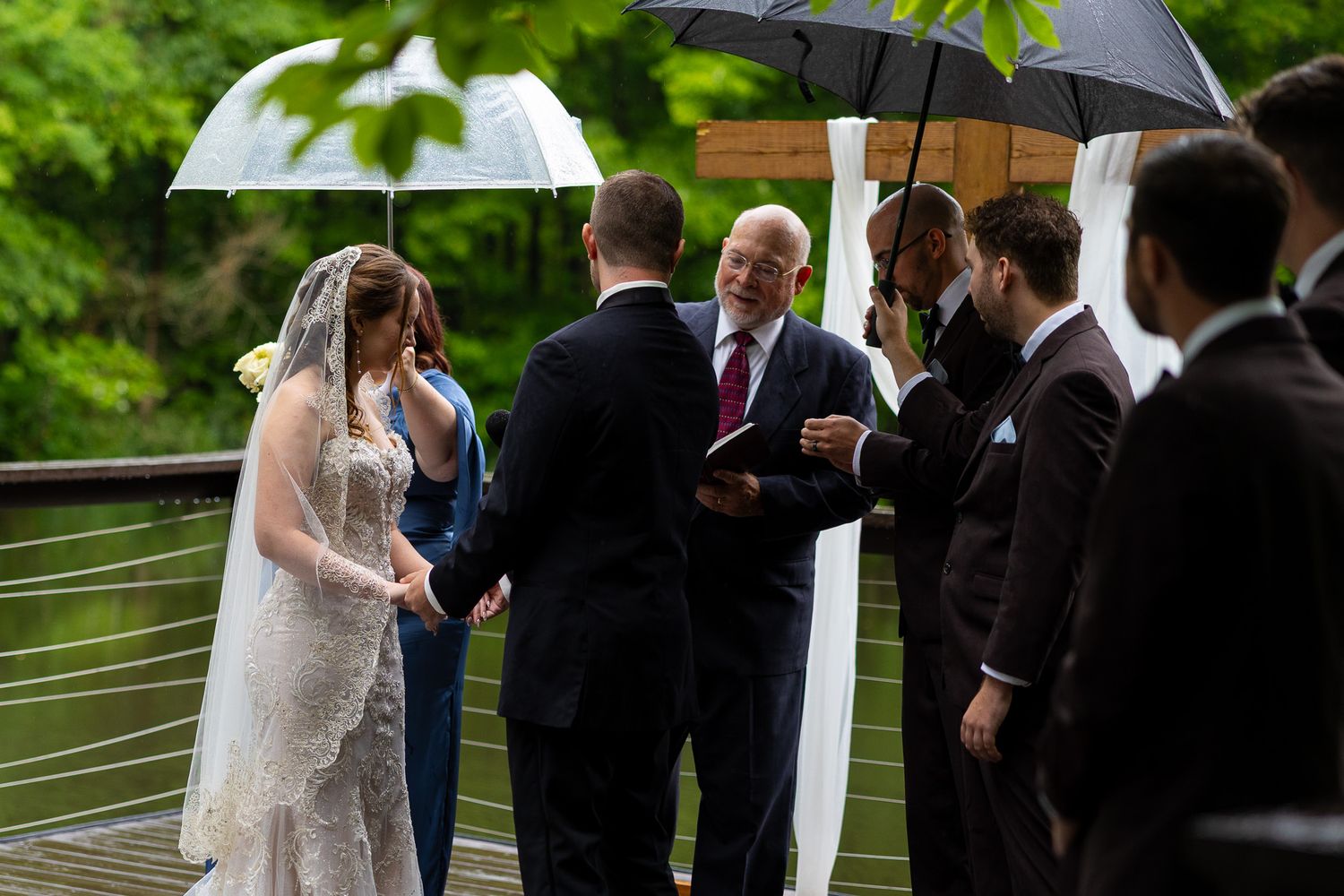 Officiant reads vows to bride and groom during intimate rainy outdoor wedding ceremony under umbrellas.