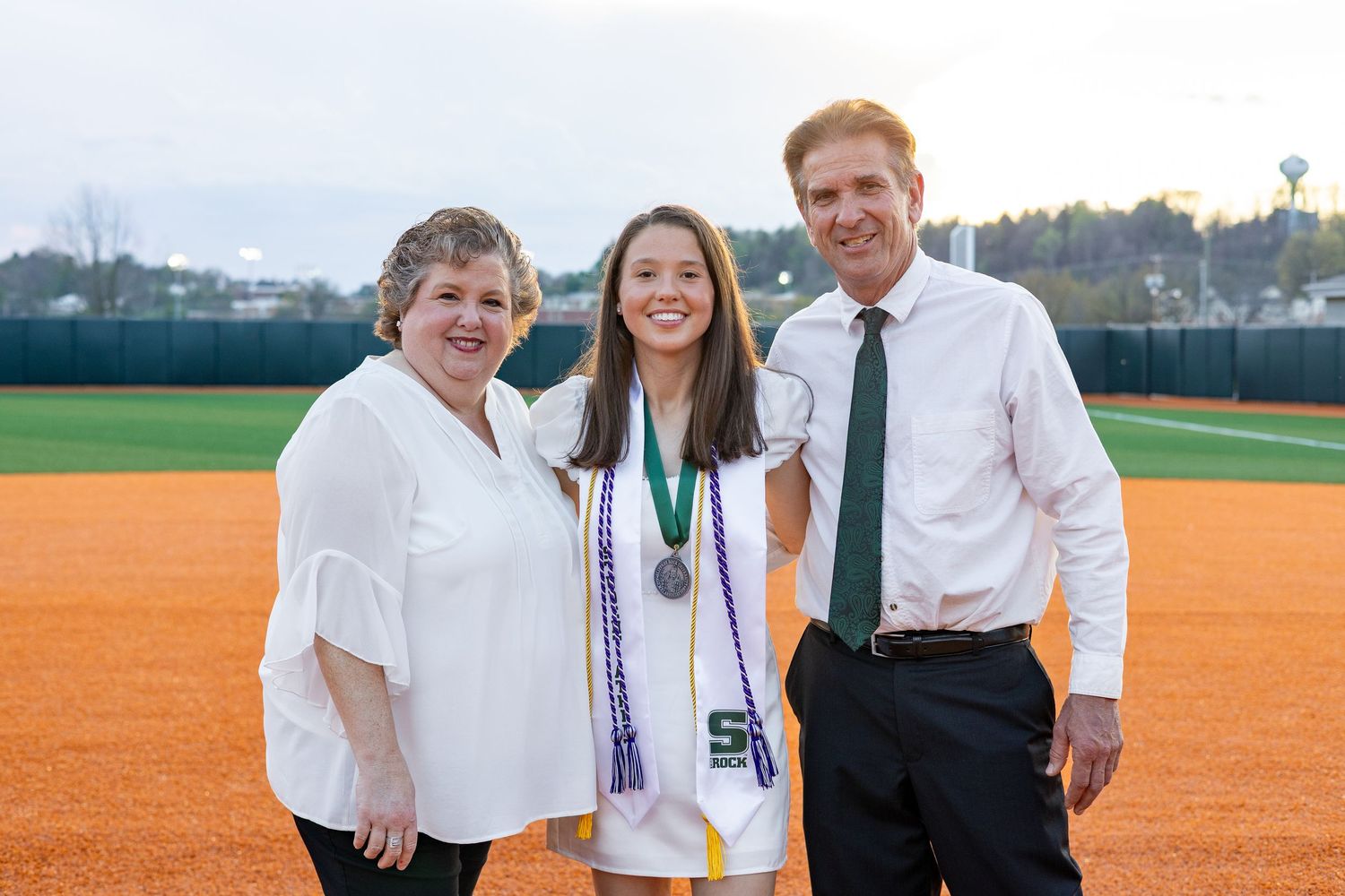 Female graduate in white dress with medal poses with parents on softball field at sunset.