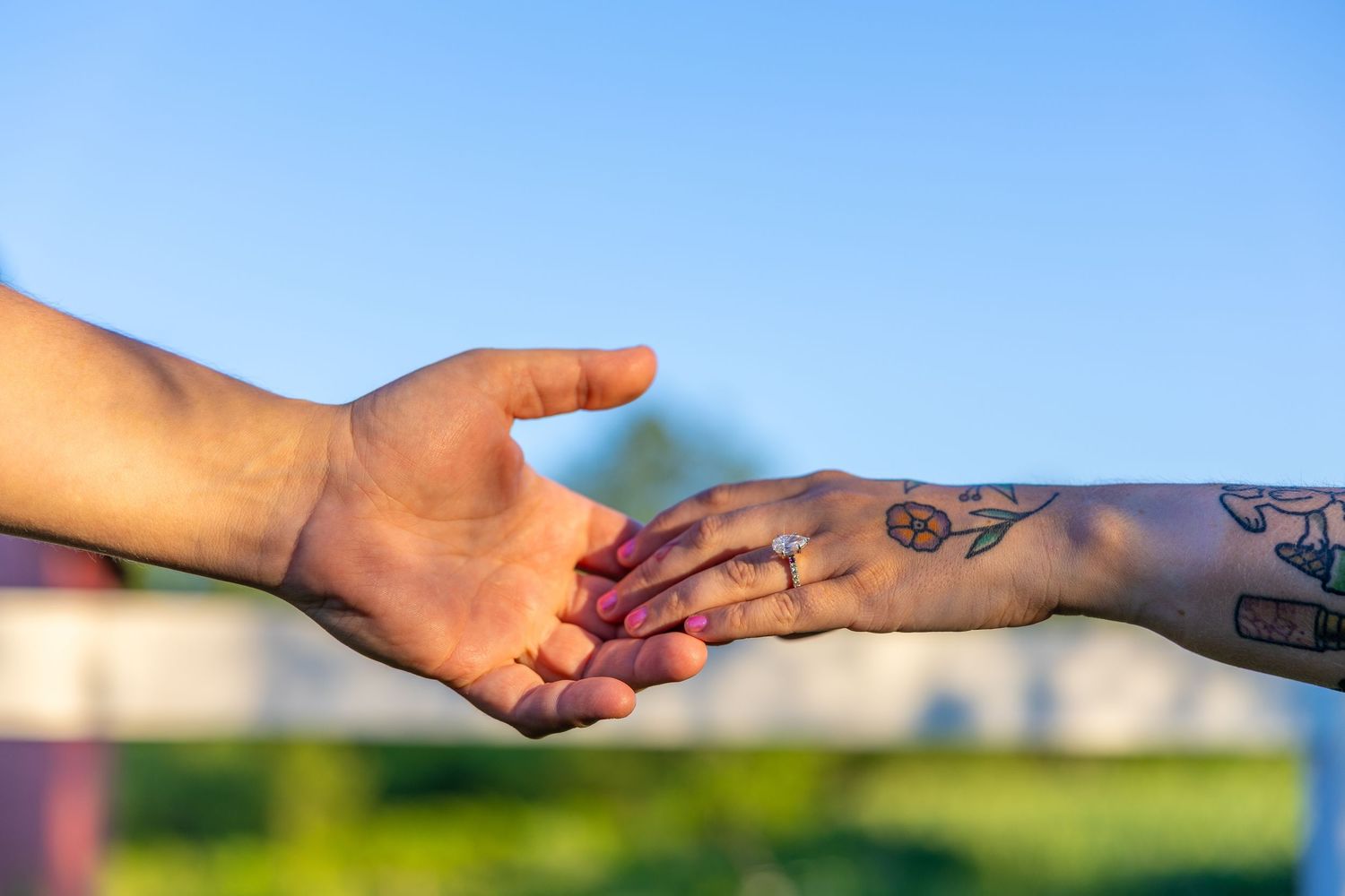 Two hands reaching toward each other, one adorned with tattoos and an engagement ring, against a blue sky background.