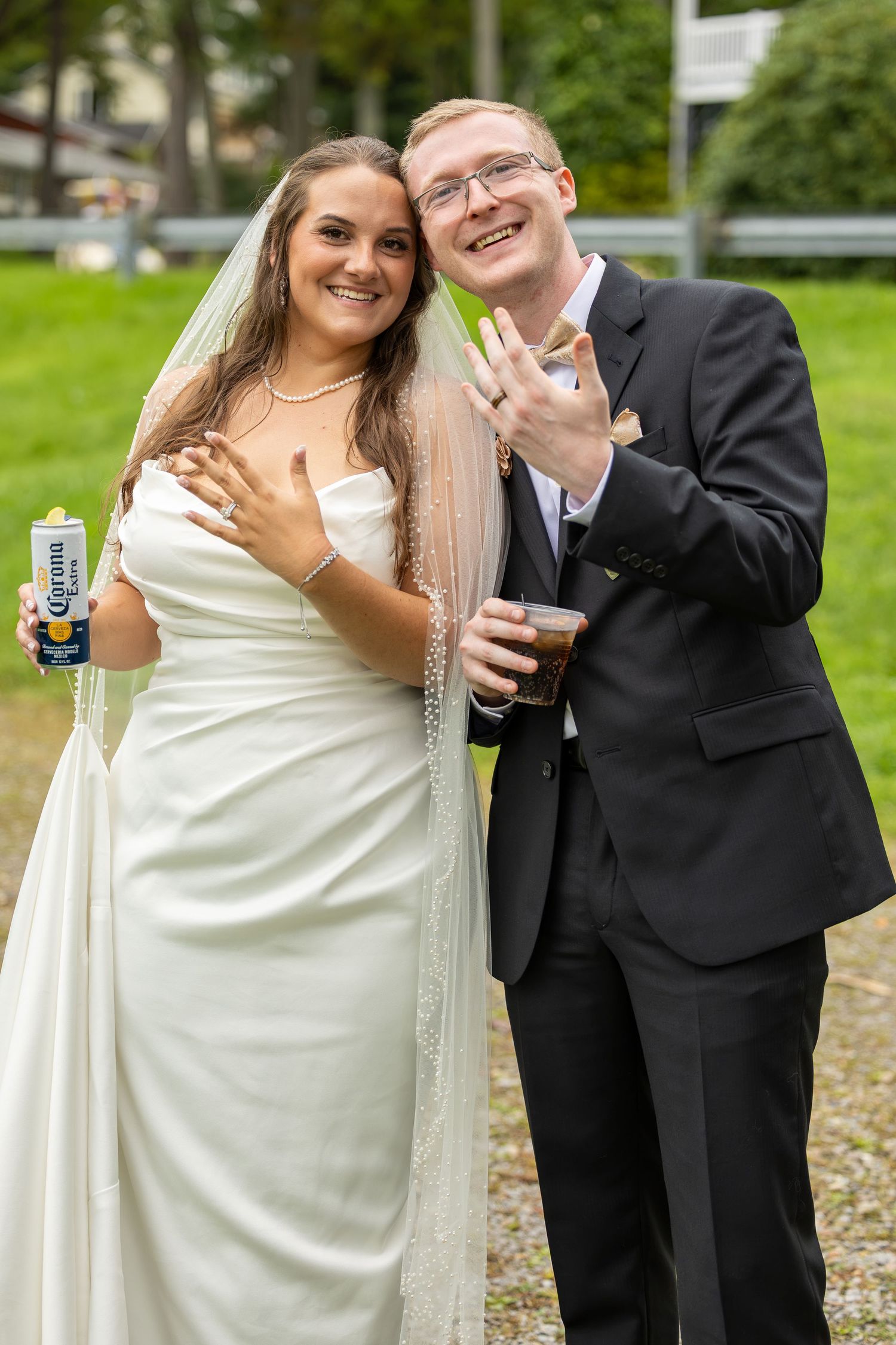 Newlywed couple posing with drinks, bride in white gown with veil holding Corona, groom in black suit showing wedding rings.