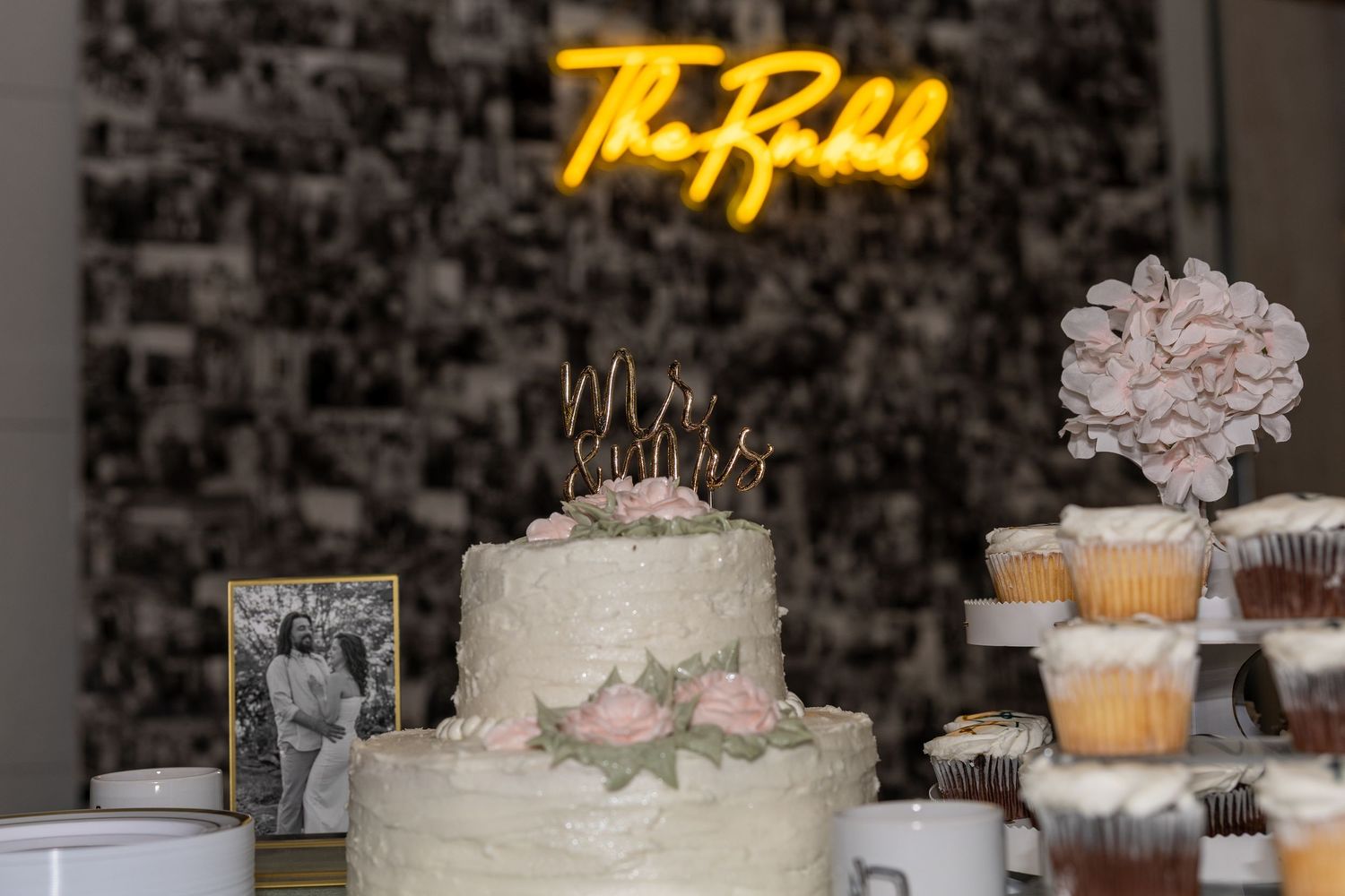 Elegant two-tier white wedding cake with pink roses and gold topper, beside cupcake tower, with neon sign backdrop.