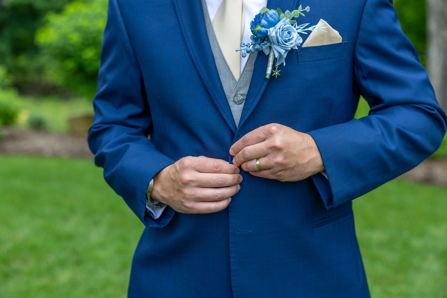 Groom in blue suit buttoning jacket with blue floral boutonniere at wedding ceremony.