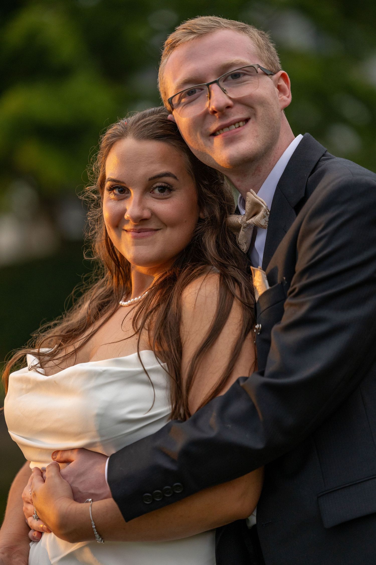 A smiling bride and groom pose together outdoors, she in a white dress, he in a gray suit with a bow tie.