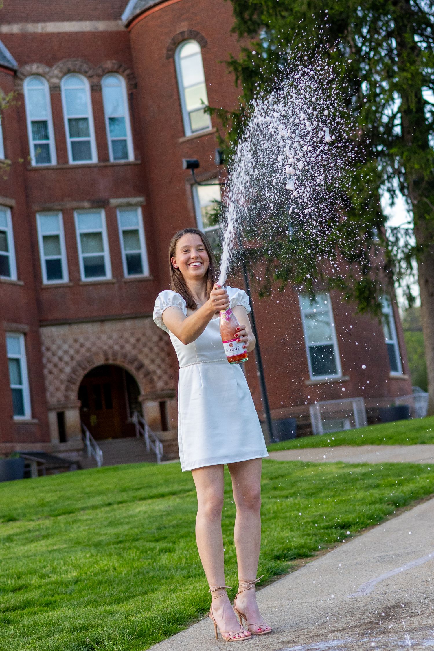 Woman in white dress celebrating graduation by popping champagne bottle outdoors in front of red brick university building.