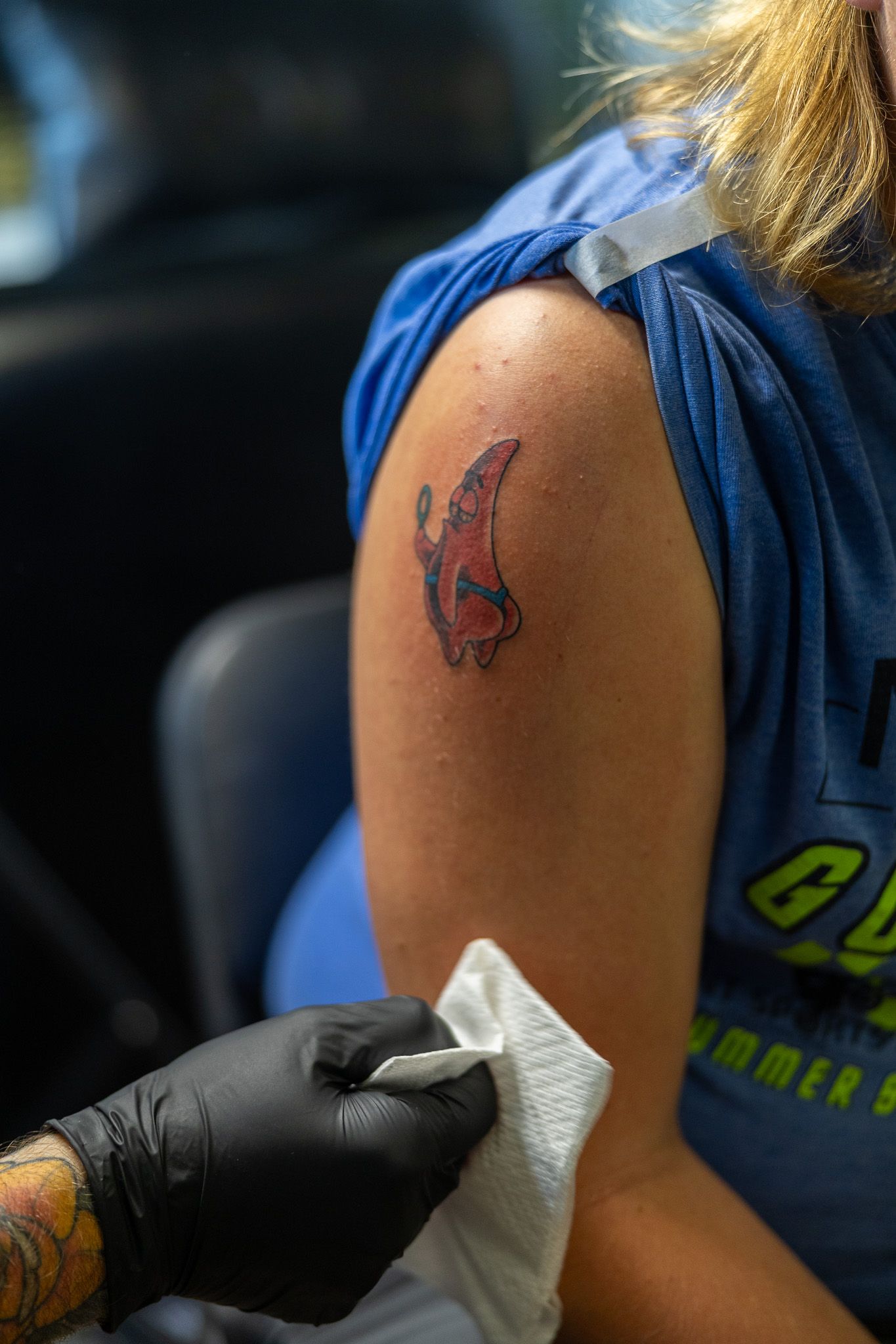 Tattoo artist wearing black gloves wiping a freshly completed red and black tattoo on a clients upper arm.