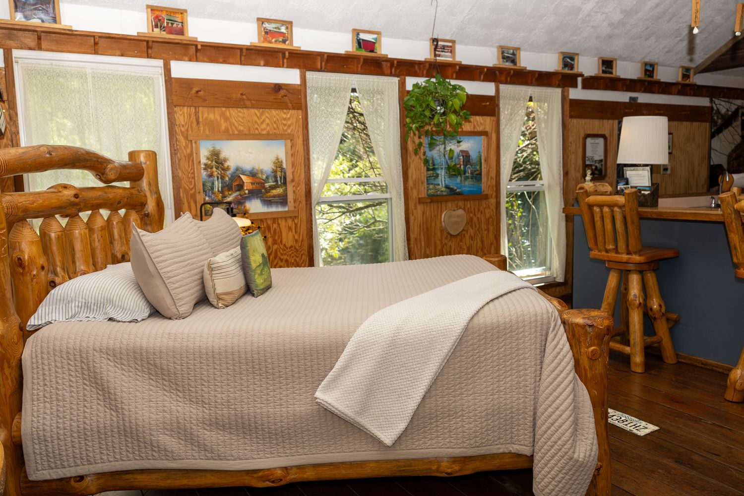 Rustic log cabin bedroom with wooden bed frame, neutral bedding, artwork on walls, and a wooden bar stool beside a desk.