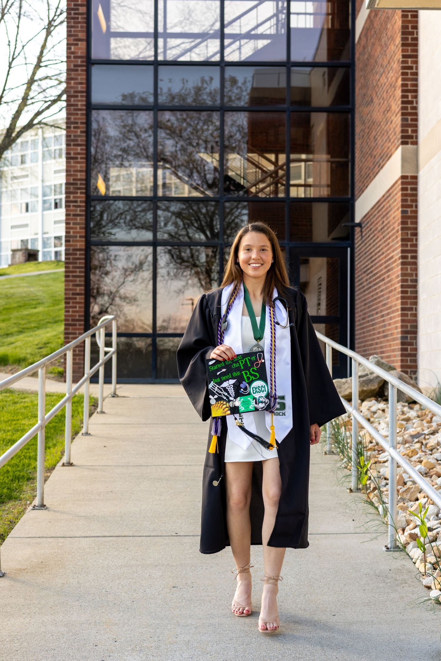 A smiling graduate in a black cap and gown walks outside a brick university building on graduation day.