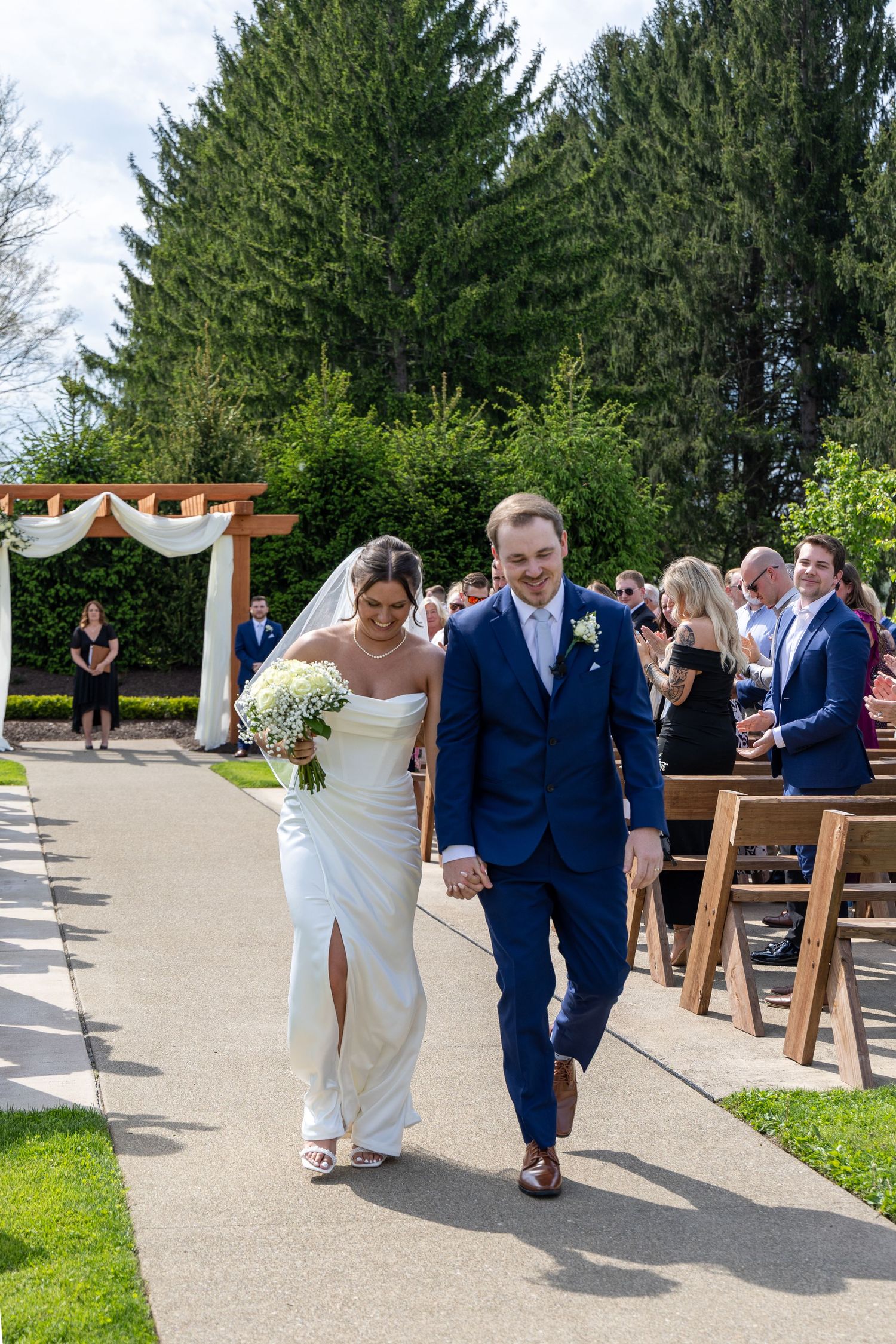 Newlyweds walk hand in hand down the aisle after outdoor wedding ceremony with guests applauding.