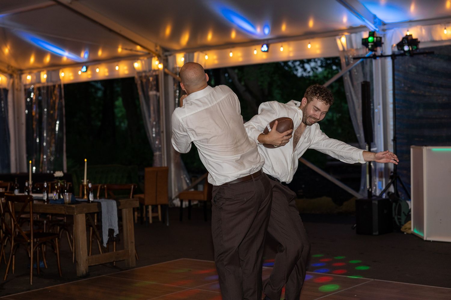 Two male wedding guests dance energetically together on the reception dance floor under string lights.