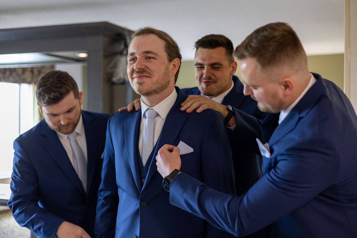 Groomsmen in navy suits helping the groom adjust his jacket and pocket square before the wedding ceremony.