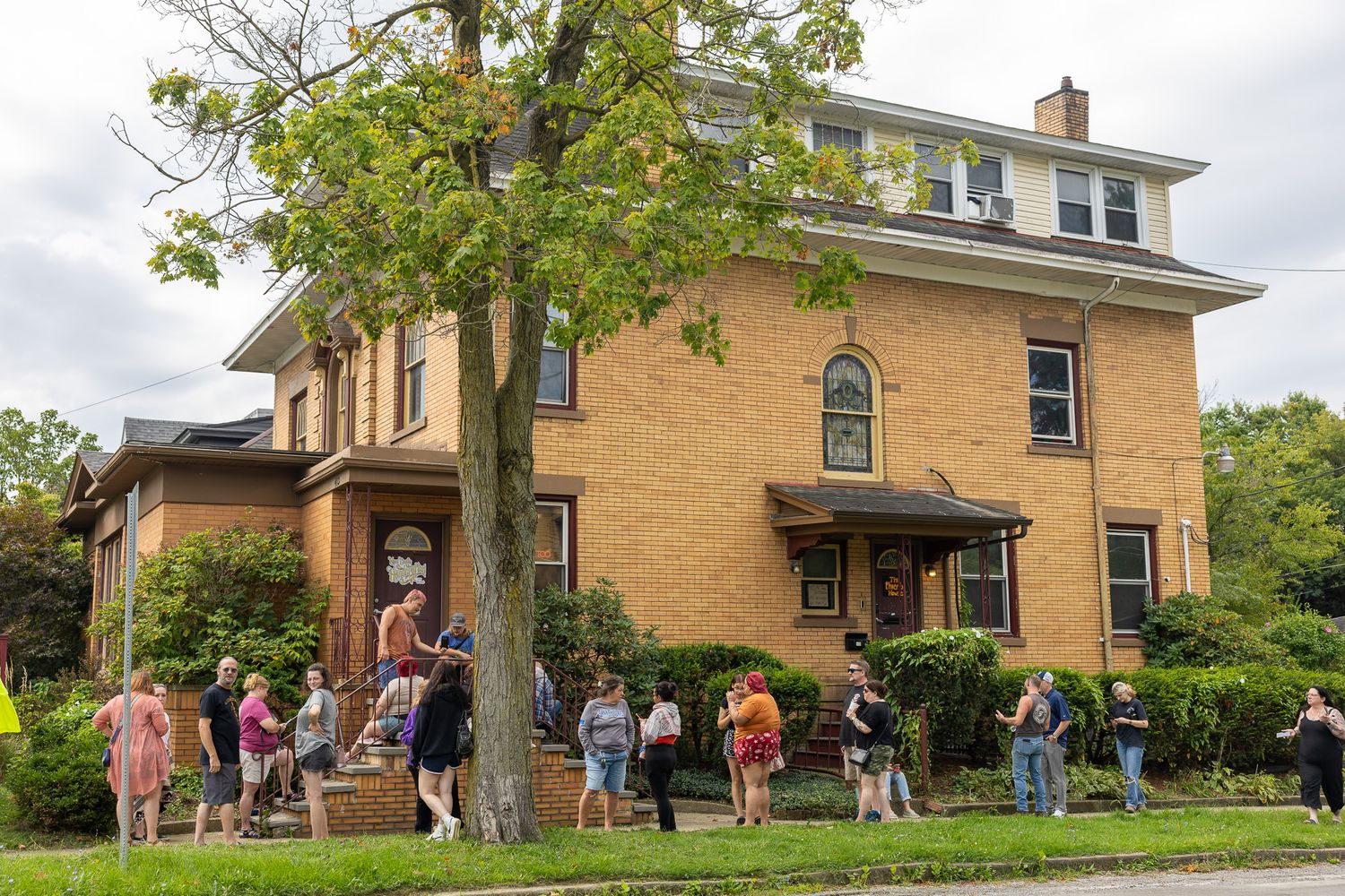 Crowd of people gathered outside a large brick two-story house with arched windows on a cloudy day.