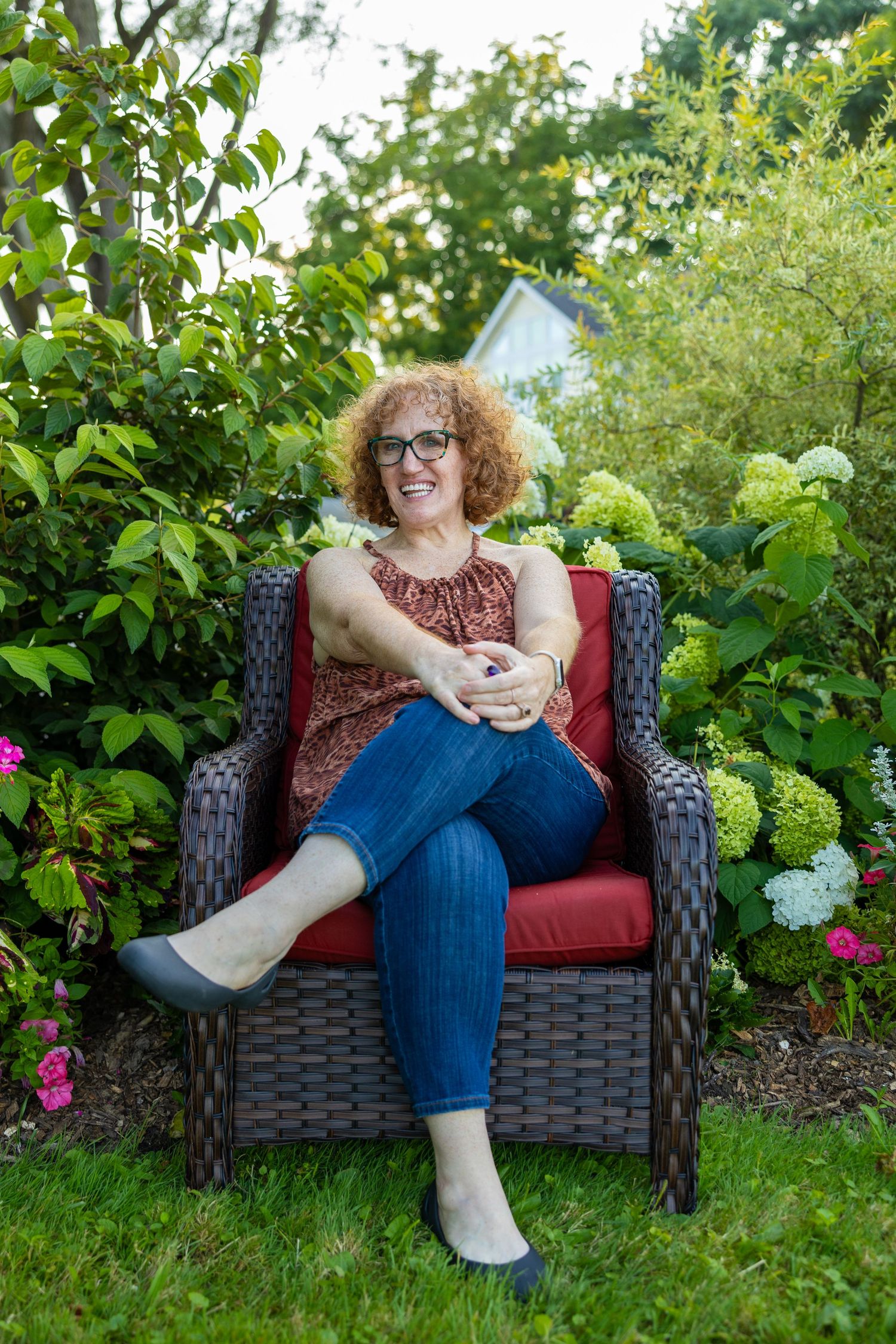 A smiling woman with curly red hair and glasses sits relaxed in a wicker chair surrounded by lush garden greenery.