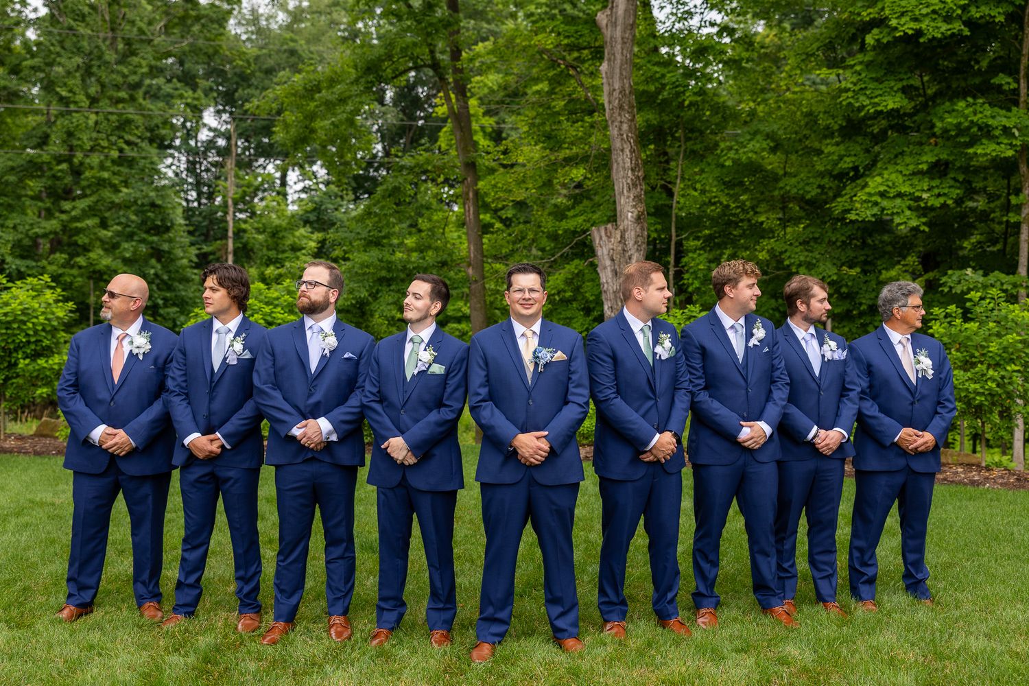 Eight groomsmen in matching navy blue suits with boutonnieres pose in a line outdoors on green grass.