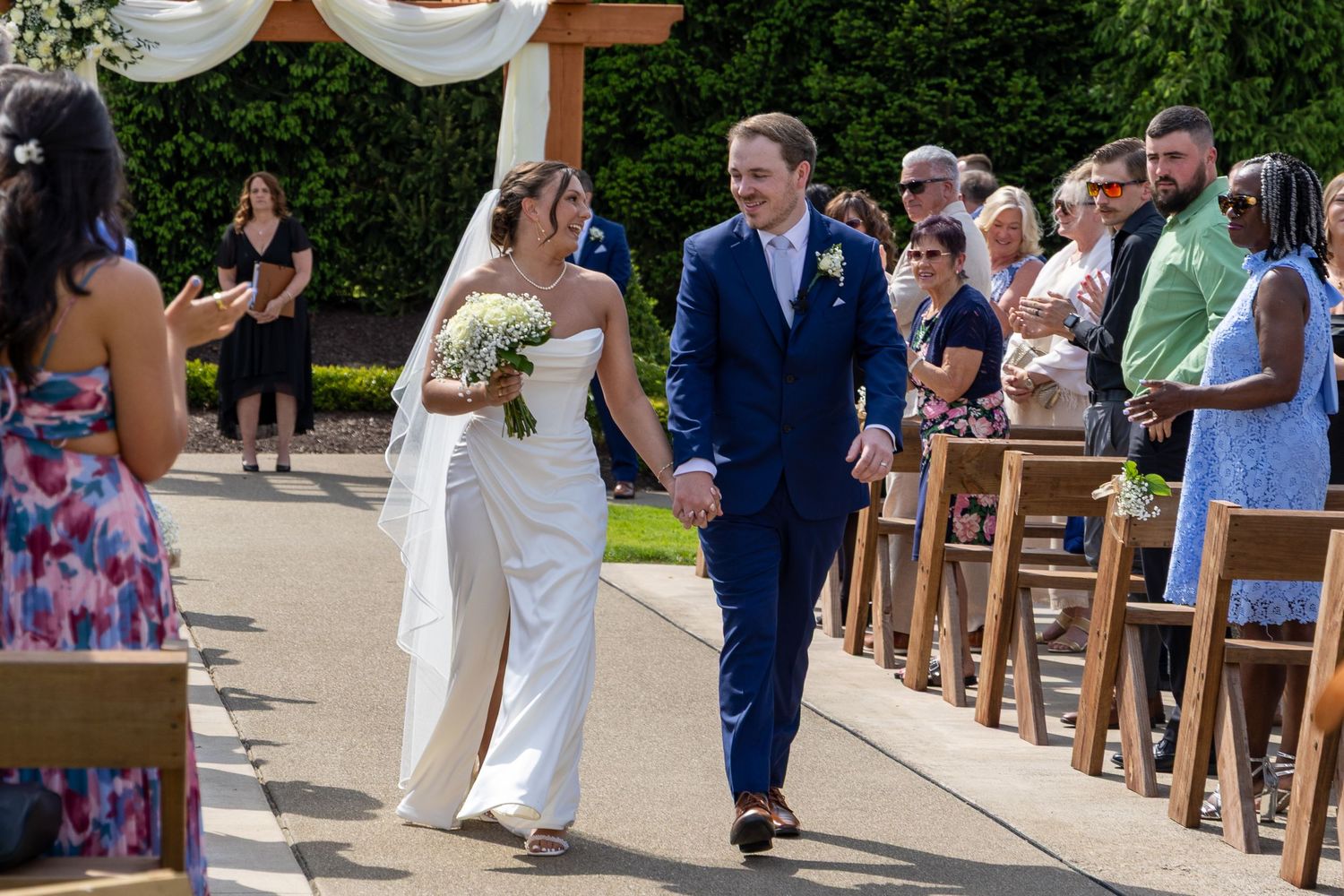 Newlywed couple walks down the aisle hand in hand after outdoor wedding ceremony with guests applauding.