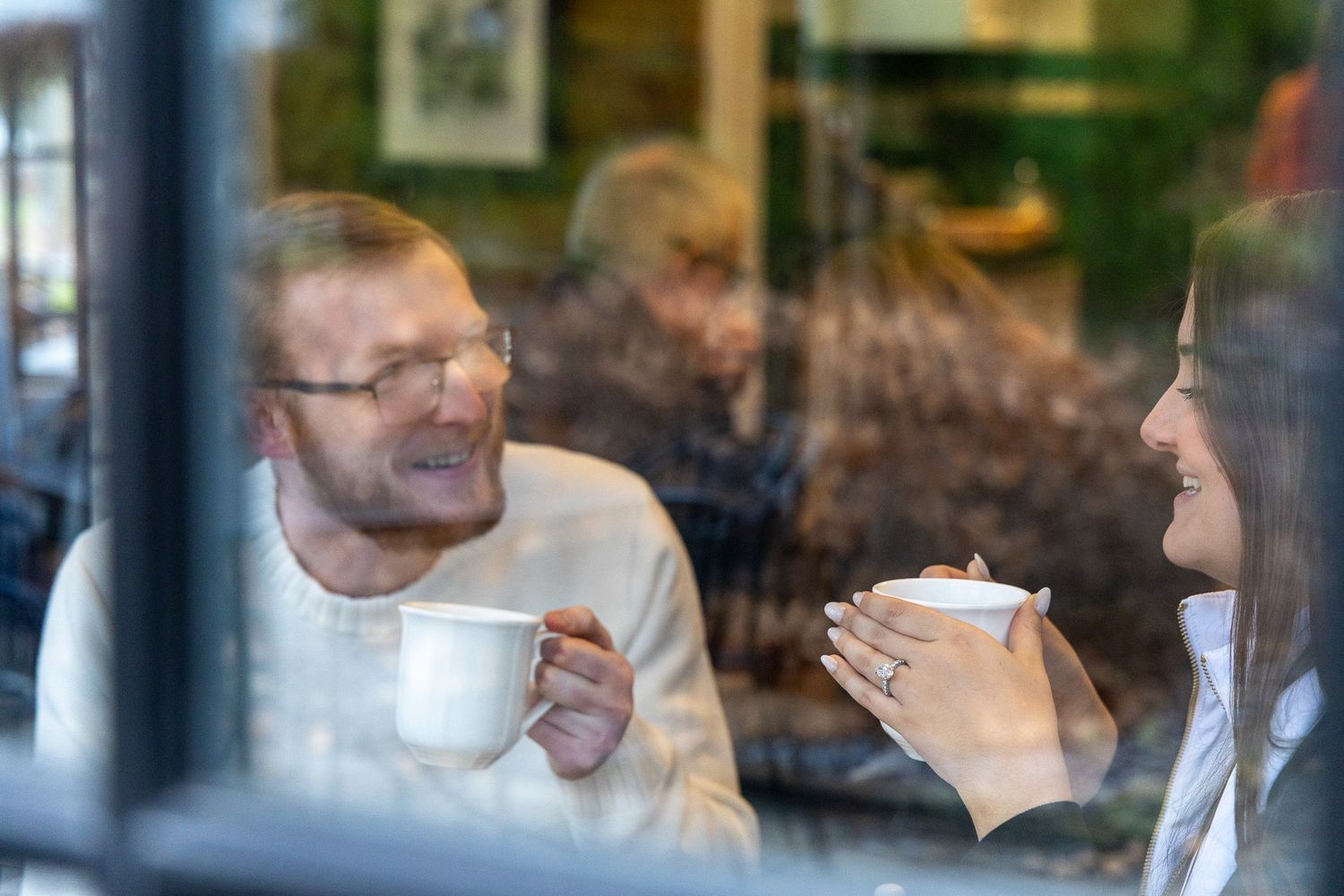 A couple enjoying coffee together at a cafe, viewed through a window with warm ambient lighting.