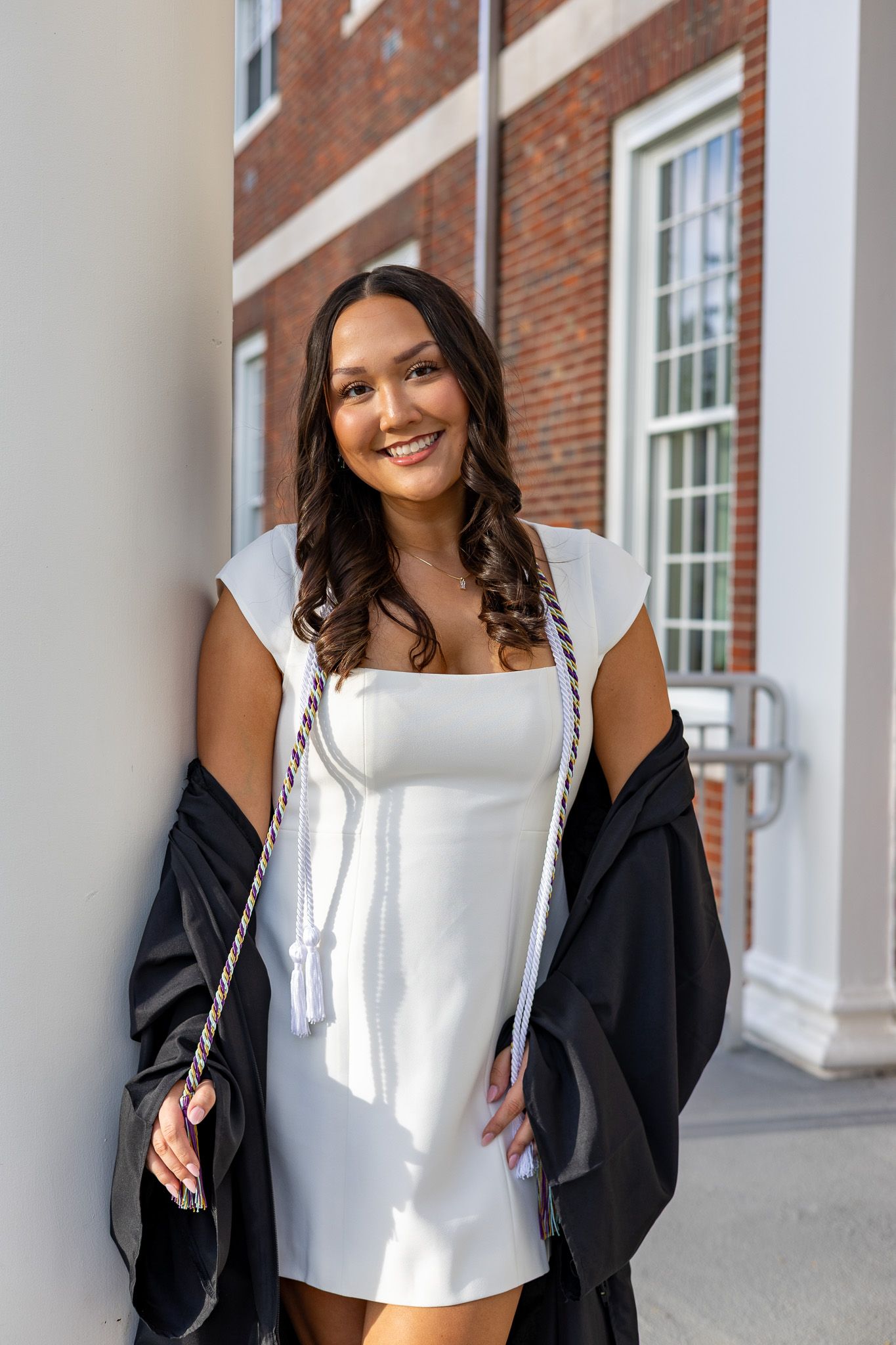 Smiling graduate in white dress holding black gown and honor cords.