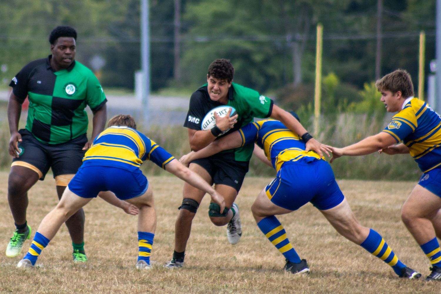 Rugby player in green and black drives through two blue and yellow defenders during a match.
