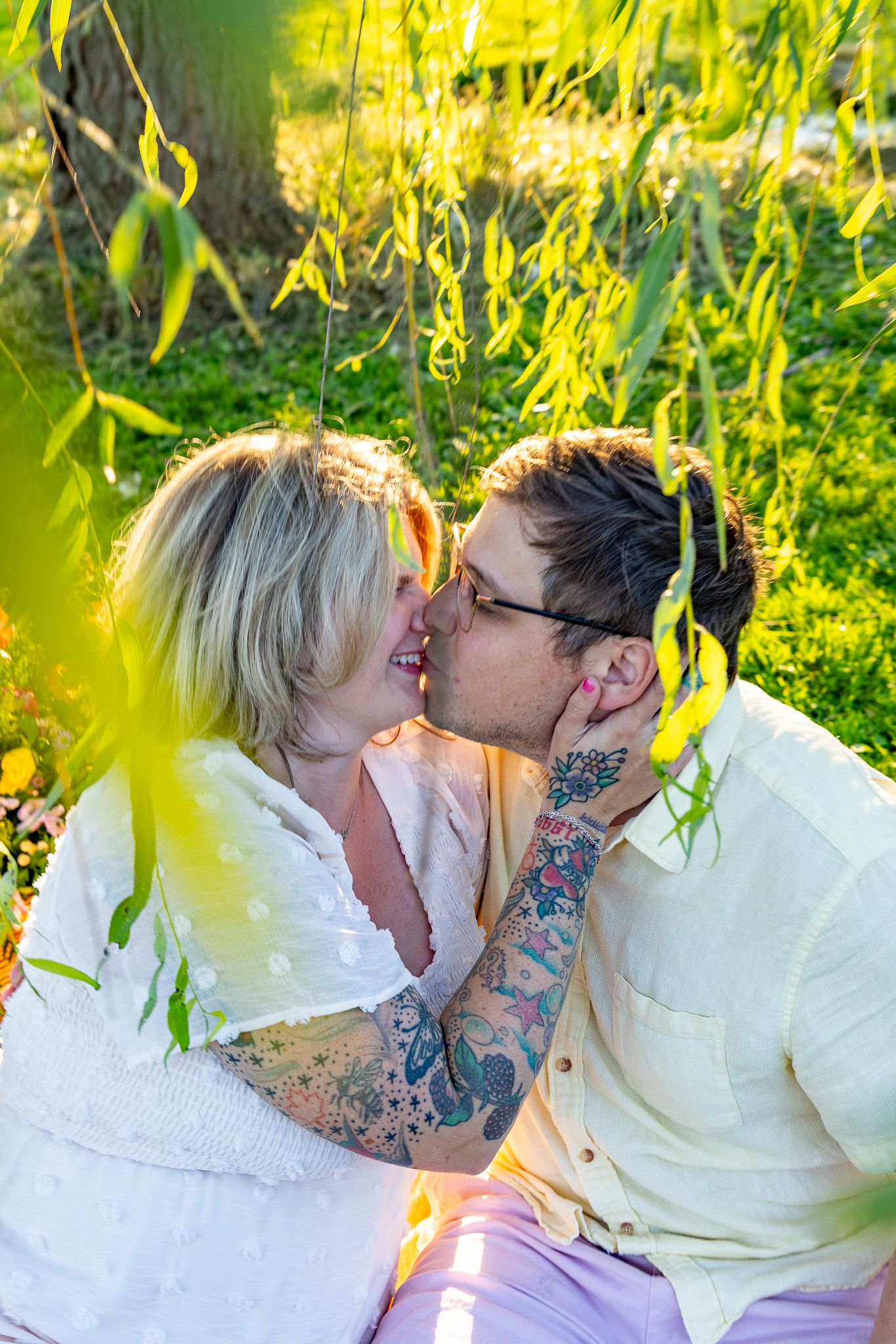 Tattooed couple sharing a kiss while sitting under a willow tree in a sunny garden.