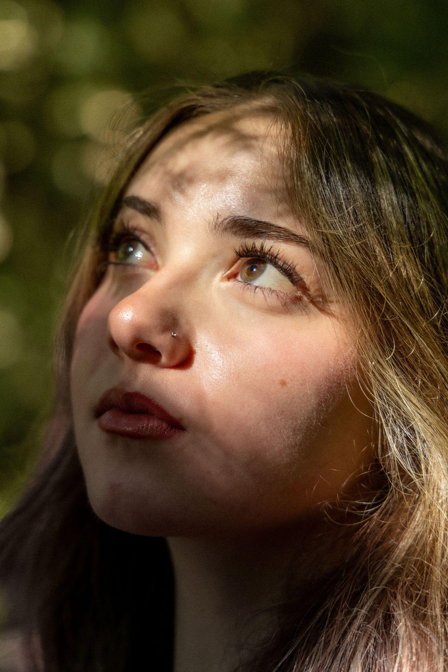 Young woman with nose ring gazing upward in dappled sunlight, with bokeh green background in close-up portrait.