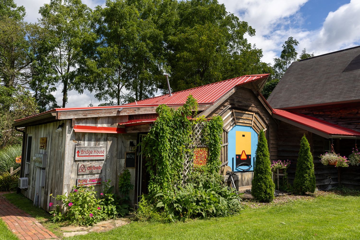 Rustic wooden shop with red metal roof, colorful murals, and lush greenery in a charming countryside setting.
