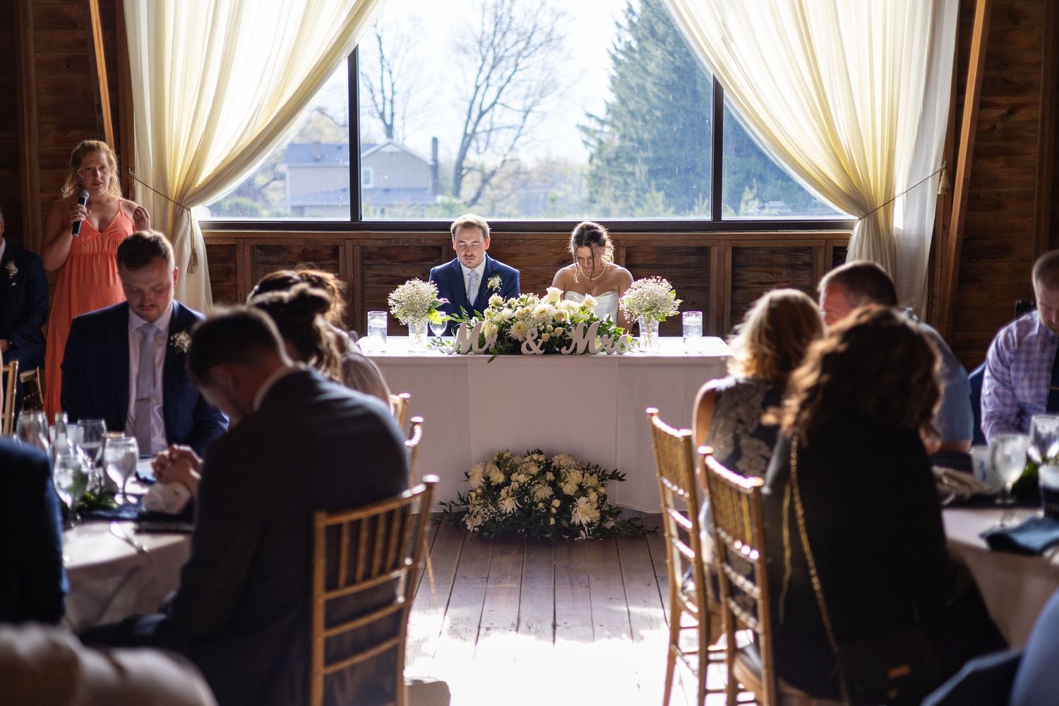 Elegant wedding reception with bride and groom at head table, surrounded by guests dining in a warmly lit venue.