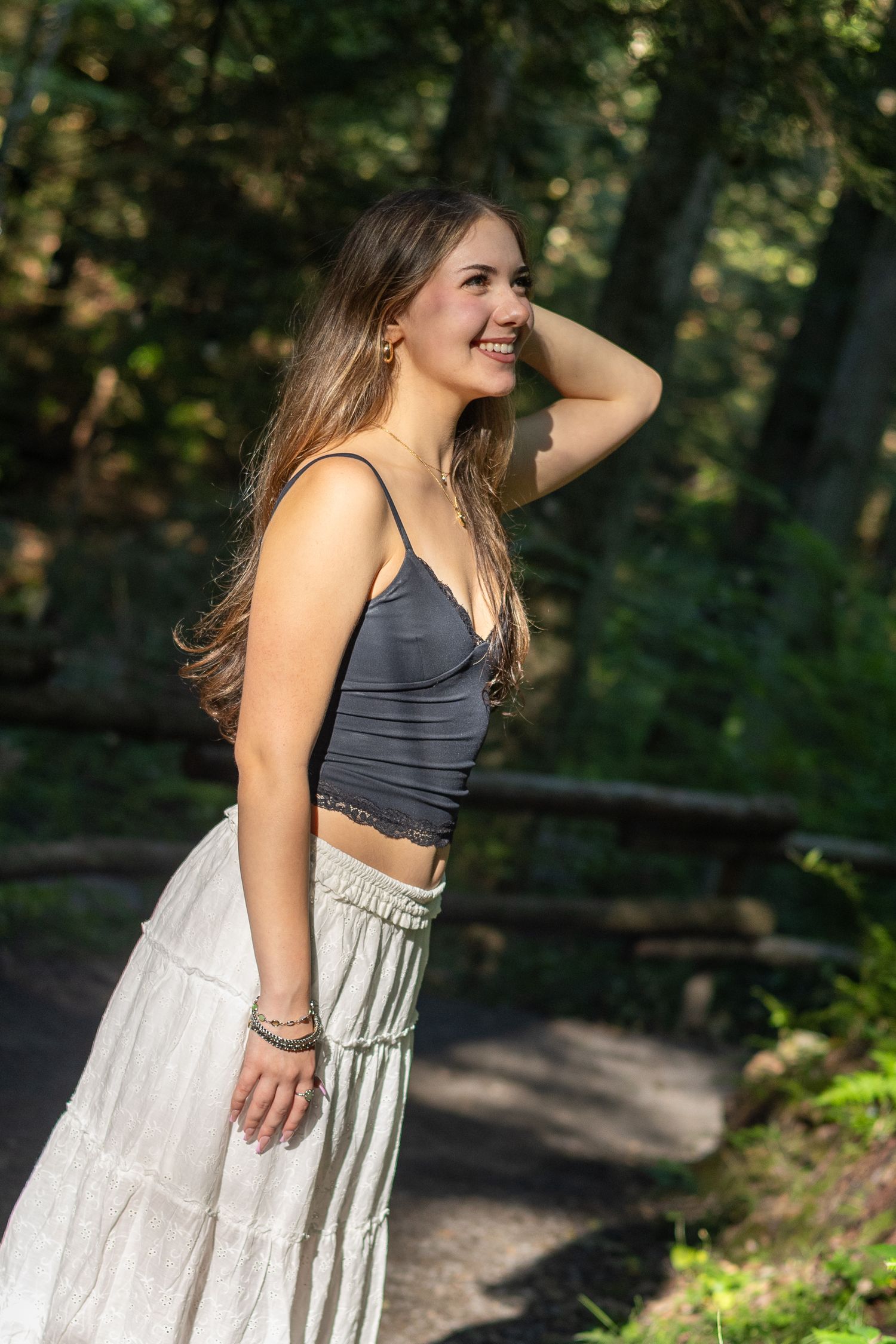 Young woman in gray crop top and flowy skirt smiling in sunlit forest path, hand in long hair.