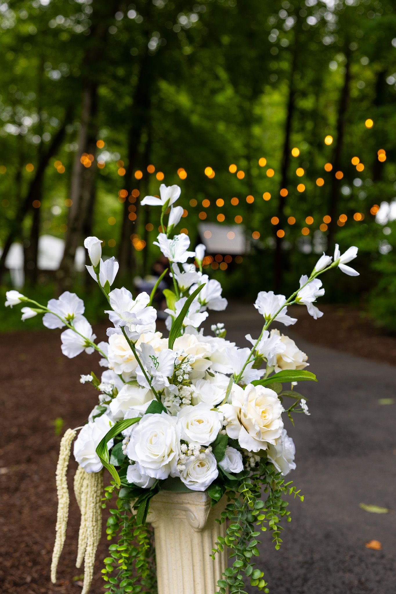 Elegant white floral arrangement with roses on a pedestal, set against a bokeh string light backdrop outdoors.