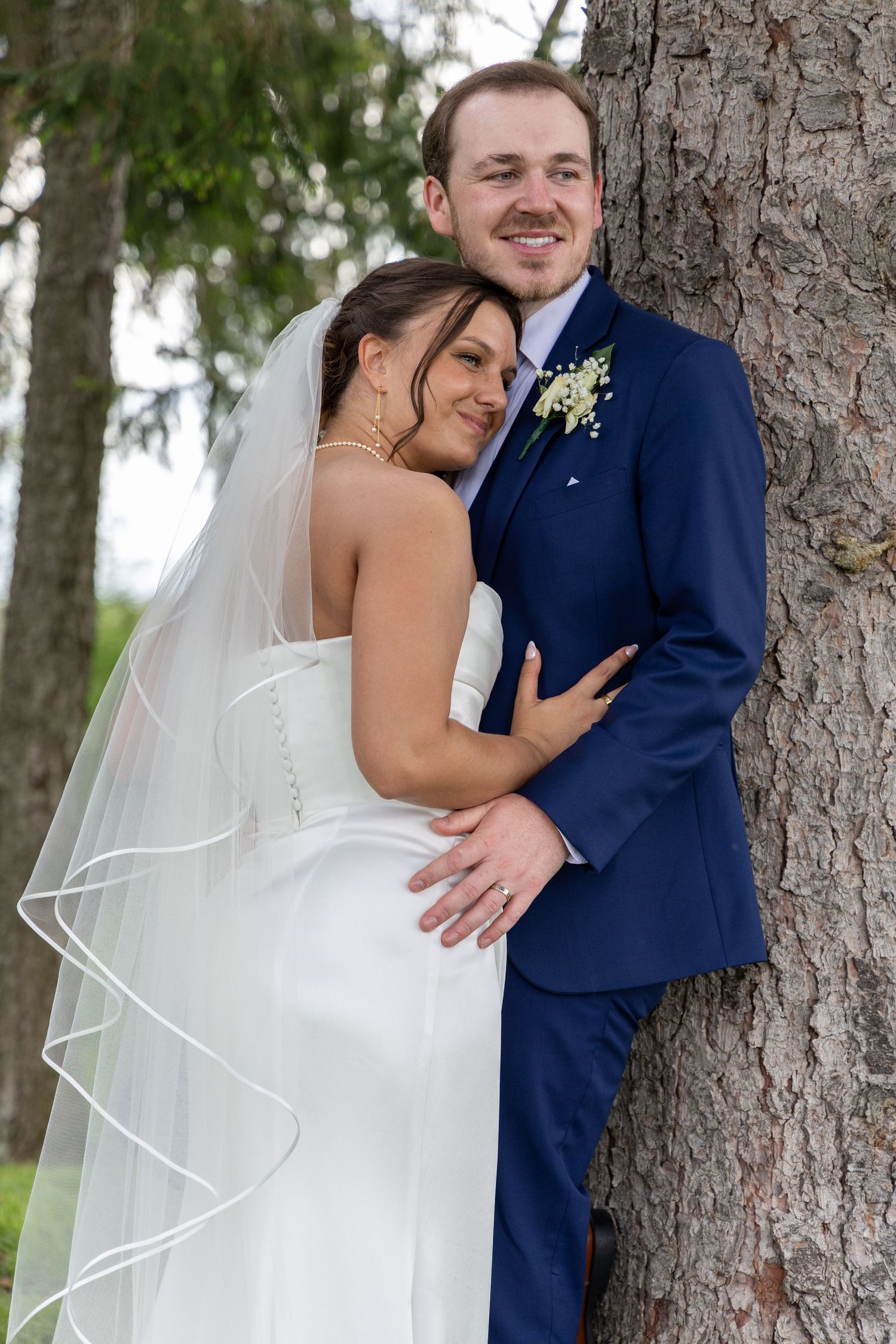 Bride in white gown and veil embraces groom in navy suit against a tree in an outdoor wedding portrait.