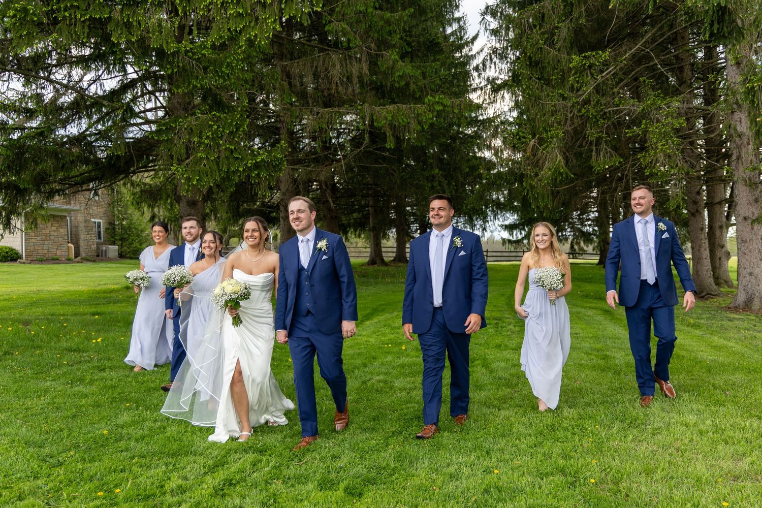 Wedding party walks across green lawn, bride in white gown with groomsmen and bridesmaids in blue.