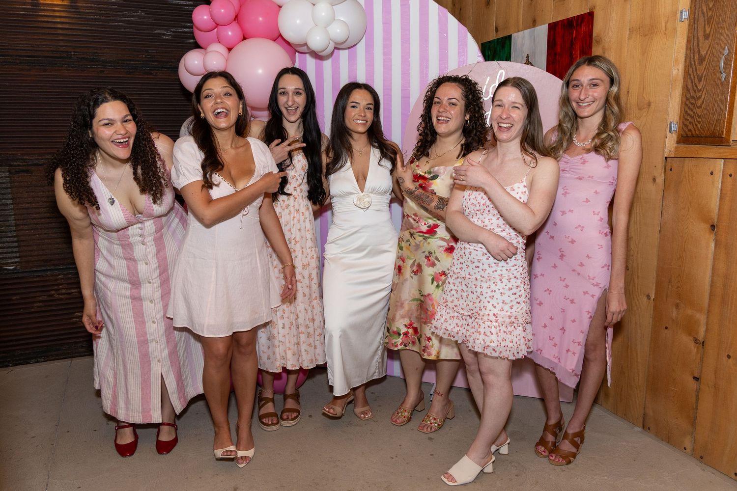 Seven women in pink and floral dresses pose together smiling at a pink balloon-decorated party backdrop.