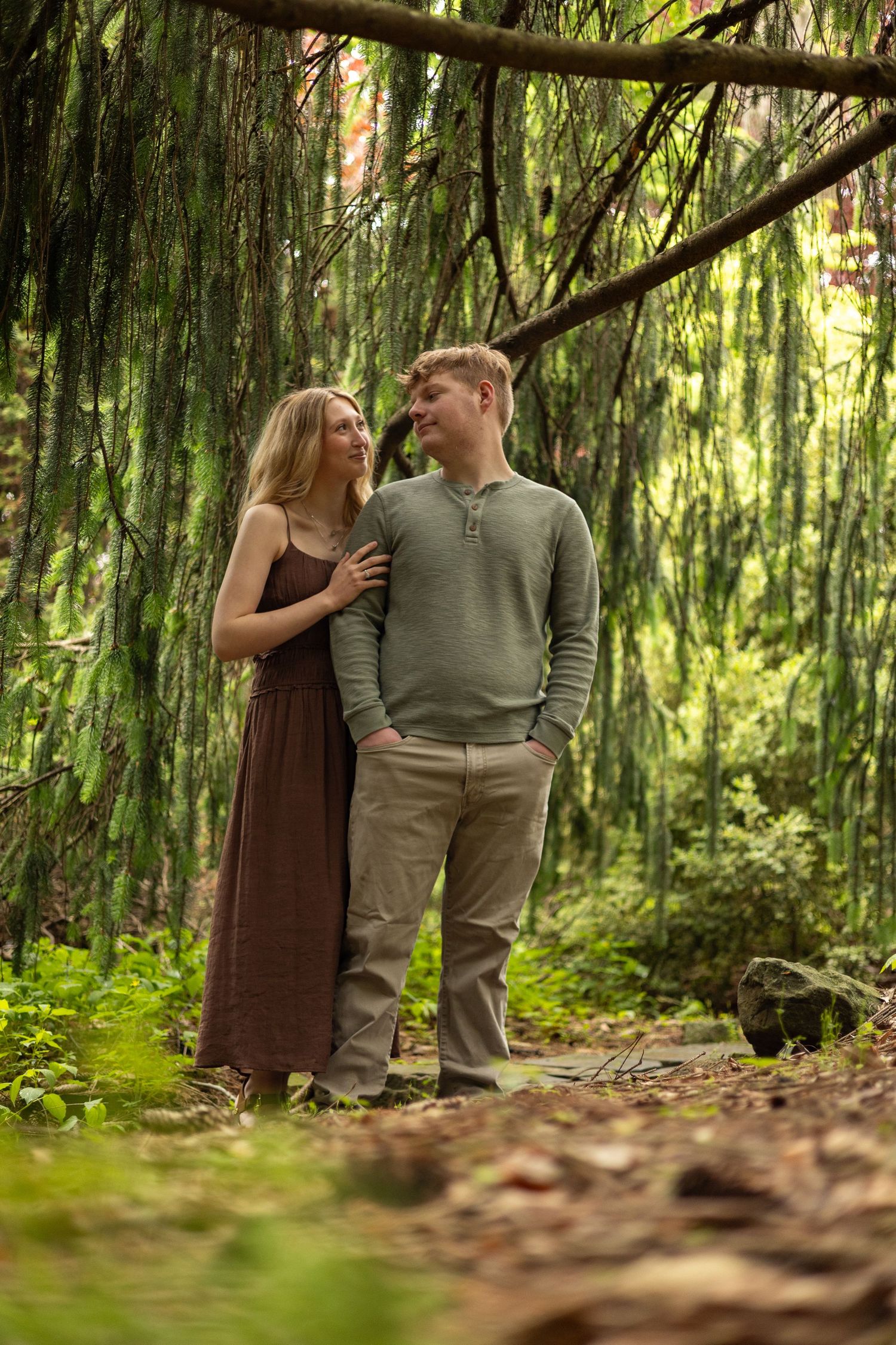 A smiling couple stands together in a lush green forest, woman resting hand on mans arm.