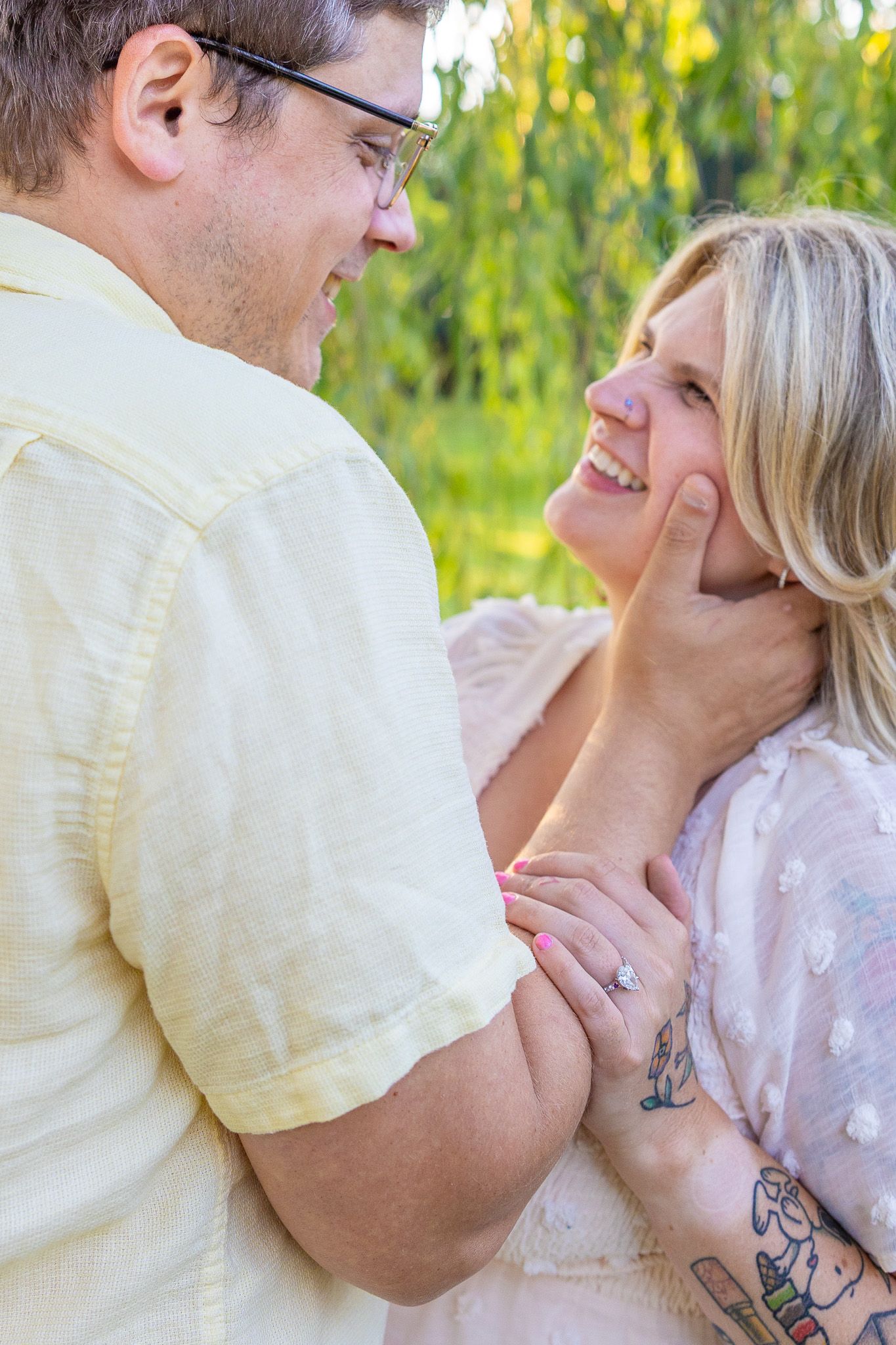 Couple gazing lovingly at each other outdoors, woman with tattoos and engagement ring smiling brightly.