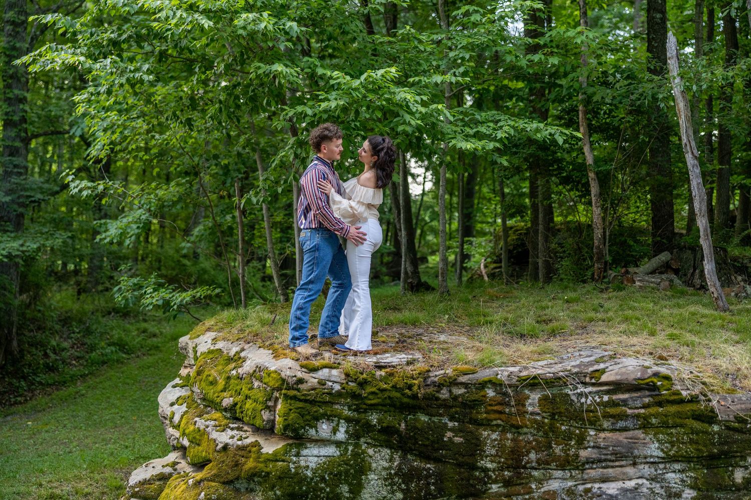 Couple standing on mossy rocks in a lush green forest, sharing a romantic moment outdoors.