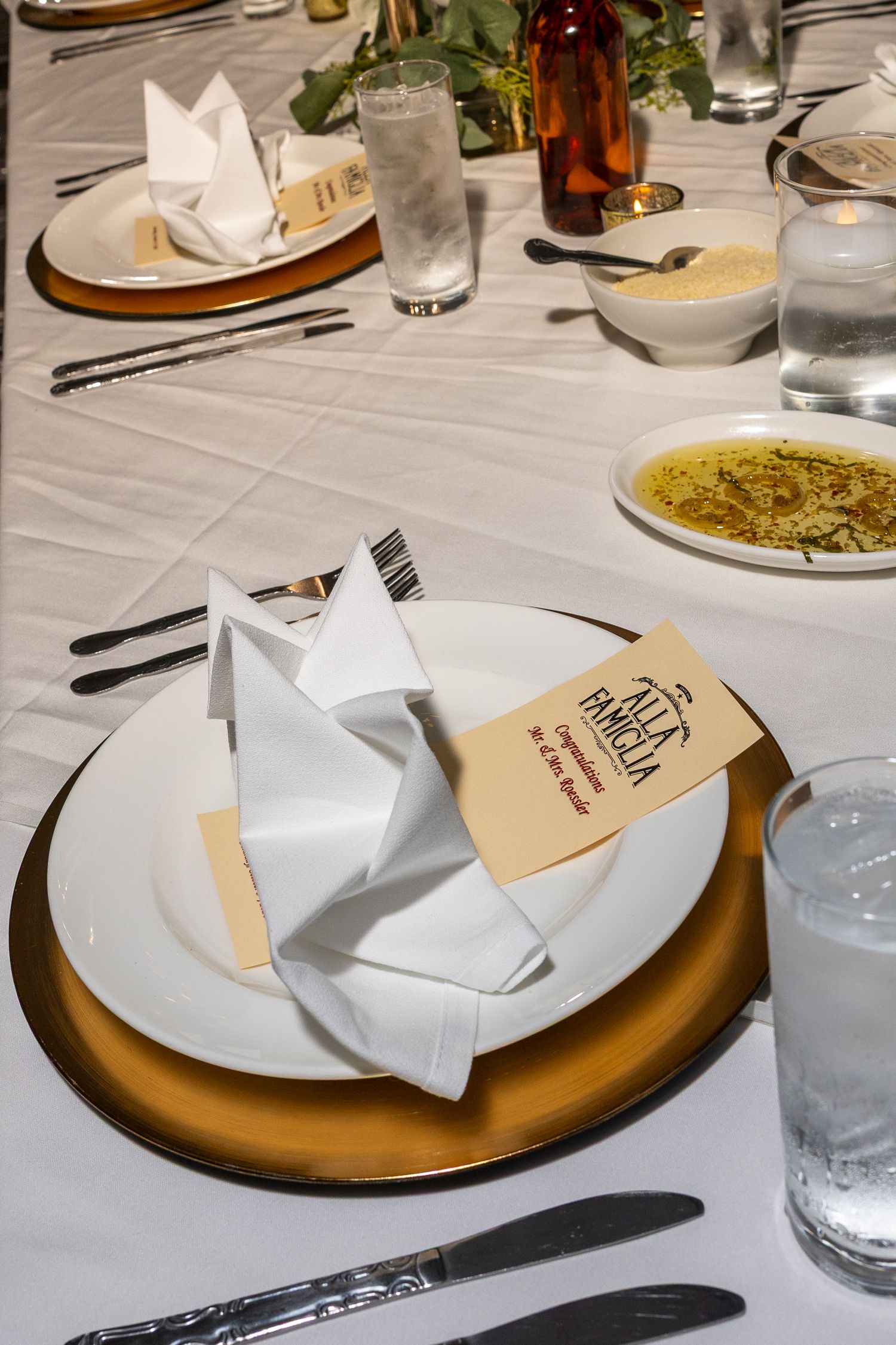 Elegant restaurant table setting with white napkin, gold charger plate, menu card, and glassware on white tablecloth.