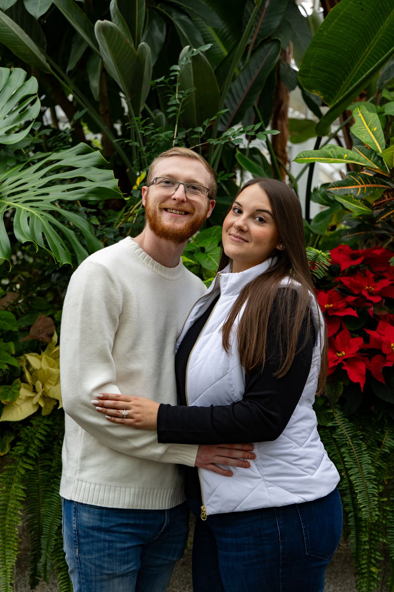 A smiling couple embraces in front of lush tropical plants and red poinsettias in a greenhouse setting.