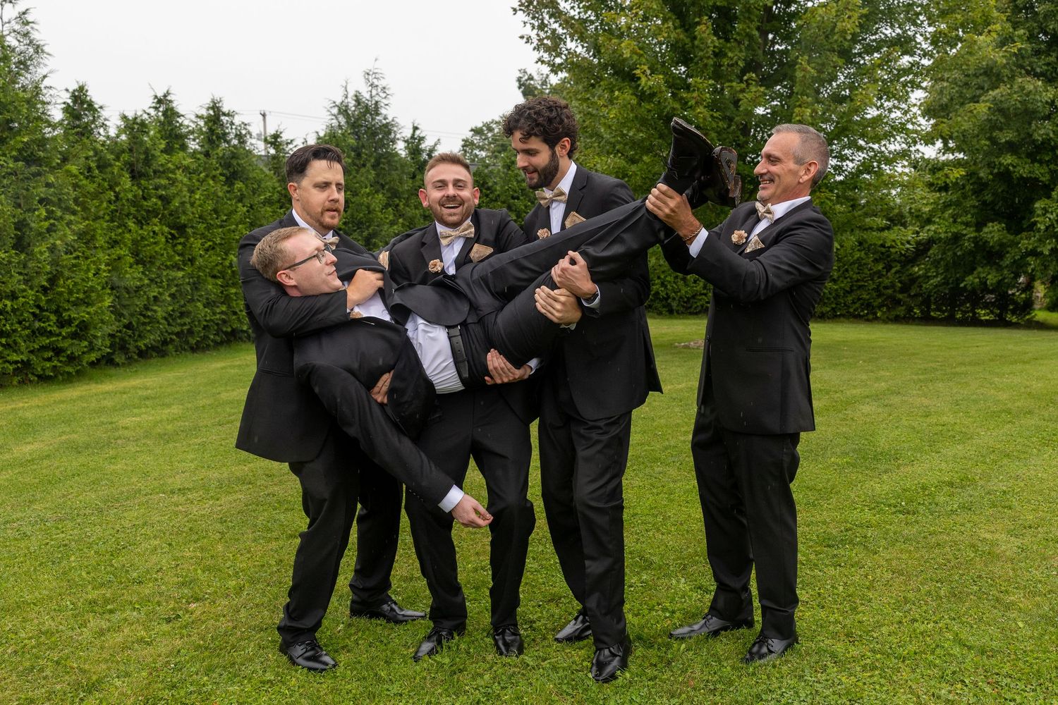 Groomsmen in black suits playfully lifting the groom outdoors on a green lawn at a wedding celebration.