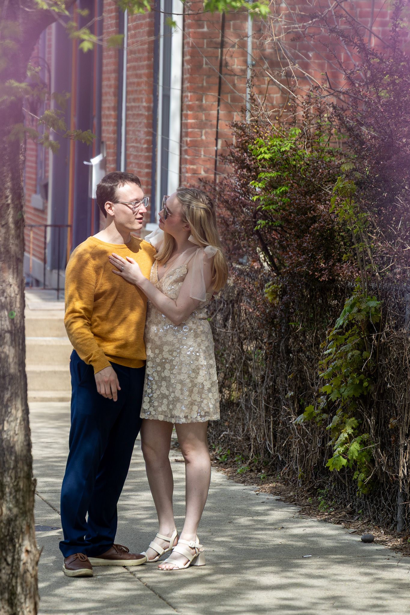 Couple sharing a romantic moment on a sunny sidewalk, man in yellow sweater and woman in floral dress, beside brick building.