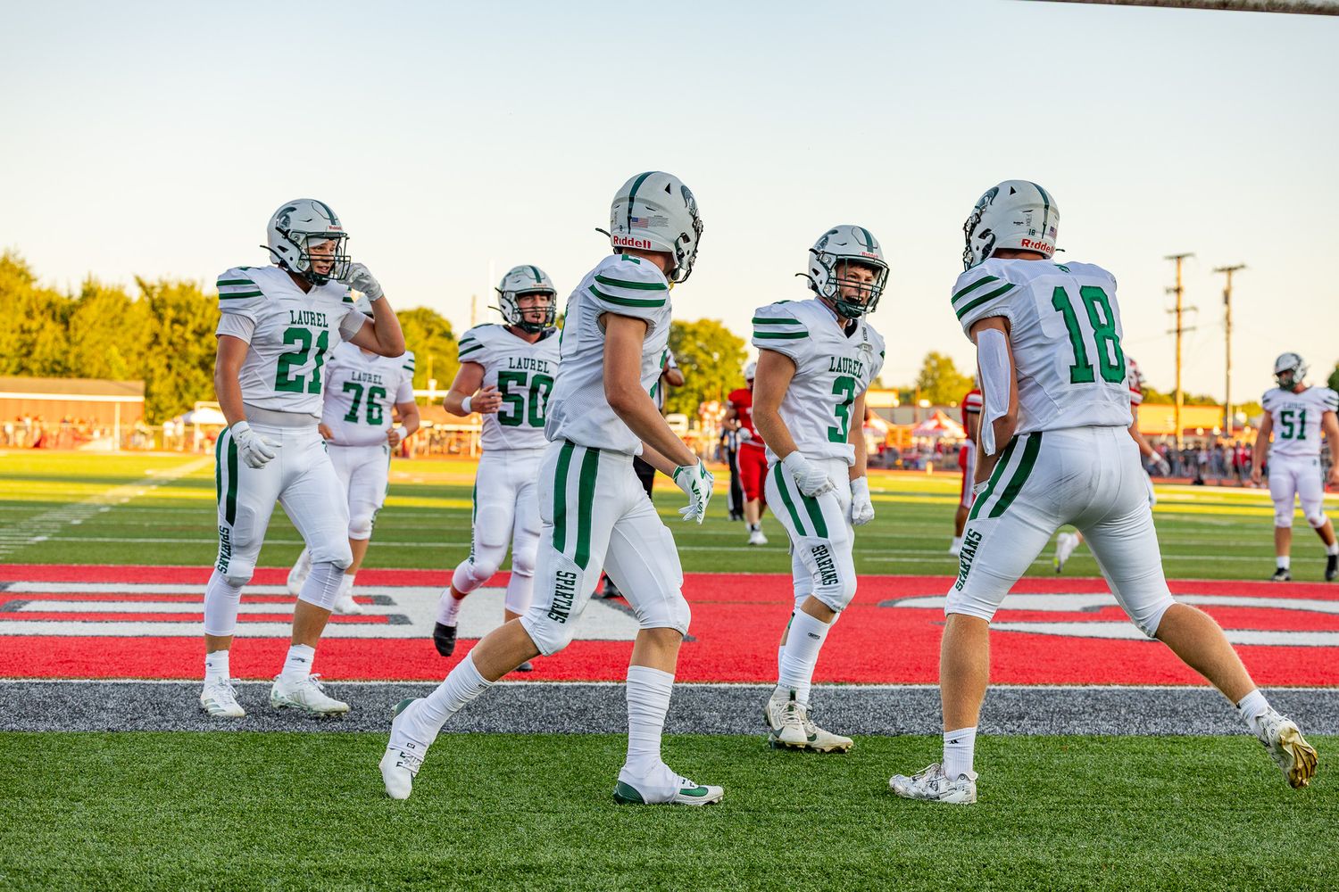 Football players in green and white uniforms walk on a red turf field during a game under warm evening light.