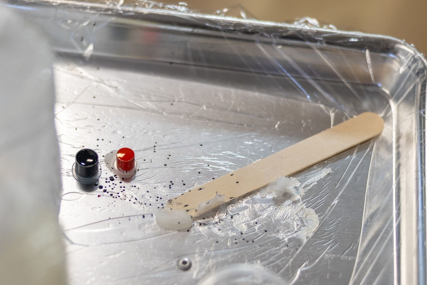 Metal tray lined with plastic wrap holding paint blobs and a wooden craft stick for mixing art supplies.