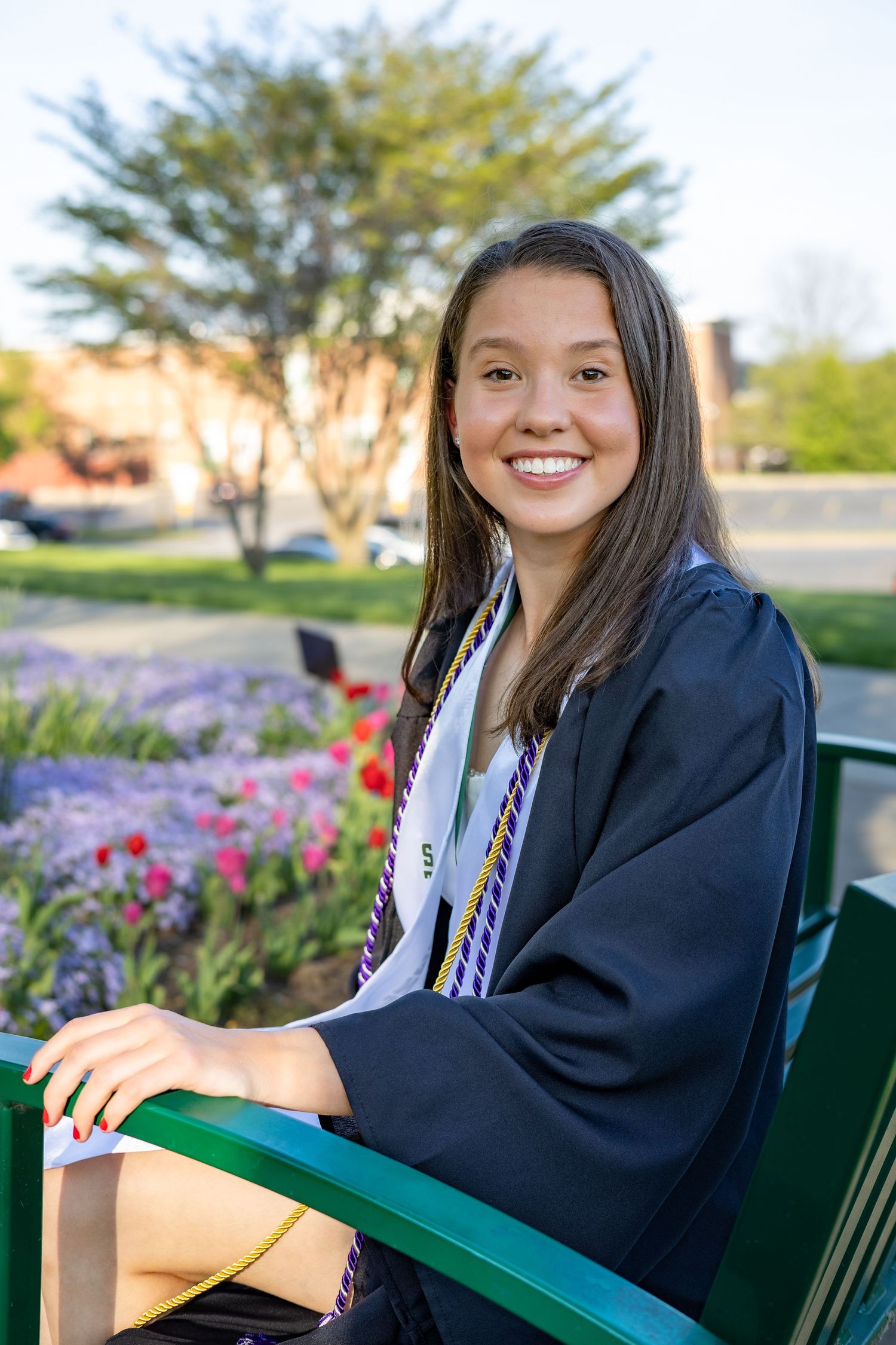Smiling graduate in cap and gown sits on green bench beside colorful spring flowers.