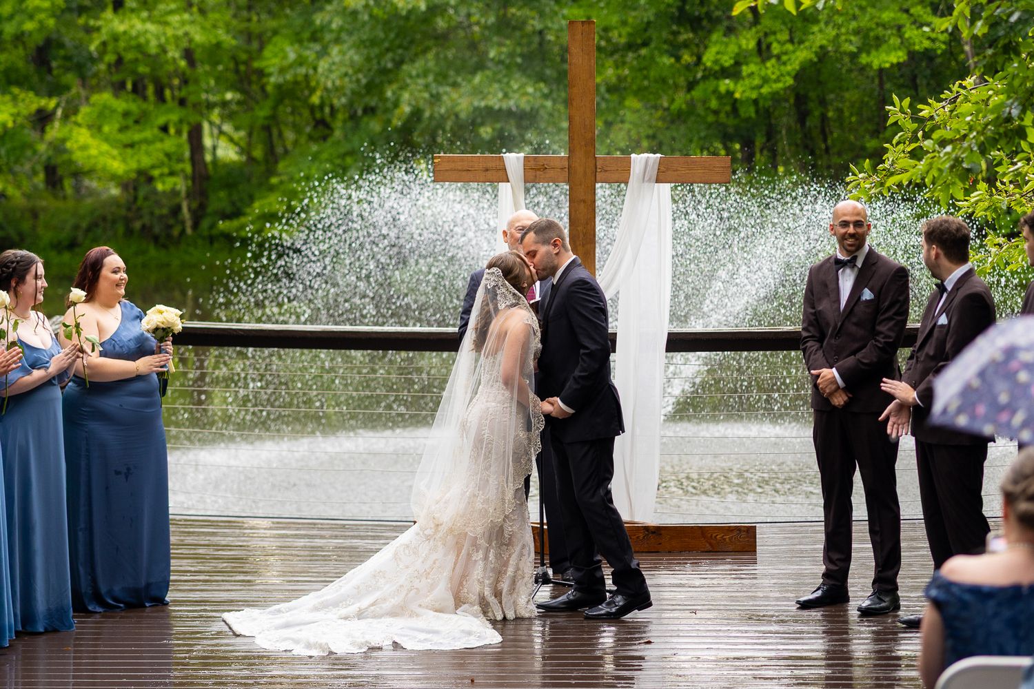 Bride and groom share first kiss at outdoor rainy wedding ceremony with cross backdrop and bridesmaids in blue dresses.