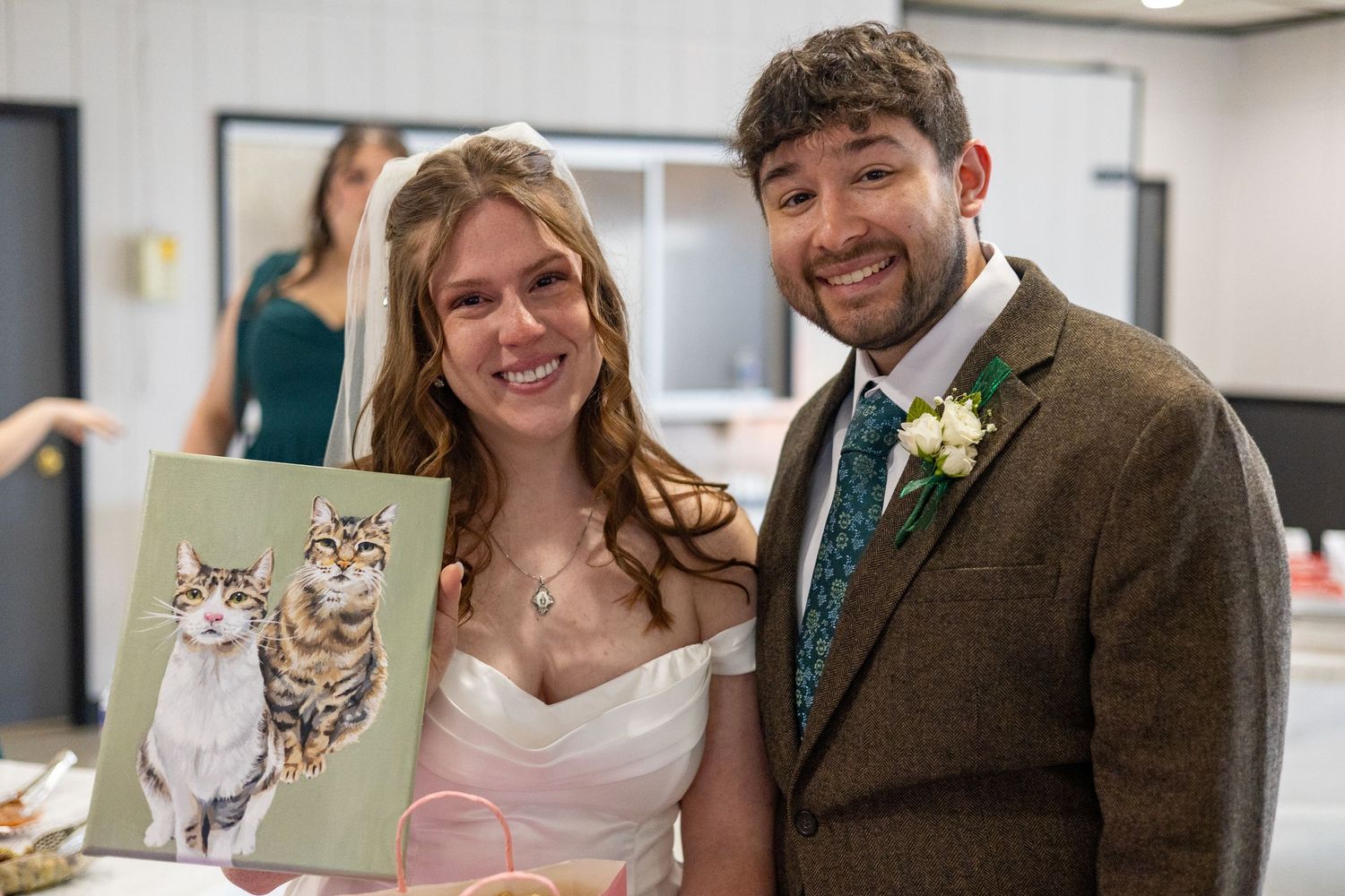 Bride and groom smiling at wedding reception, holding a painted portrait of two cats, groom wearing brown tweed suit.