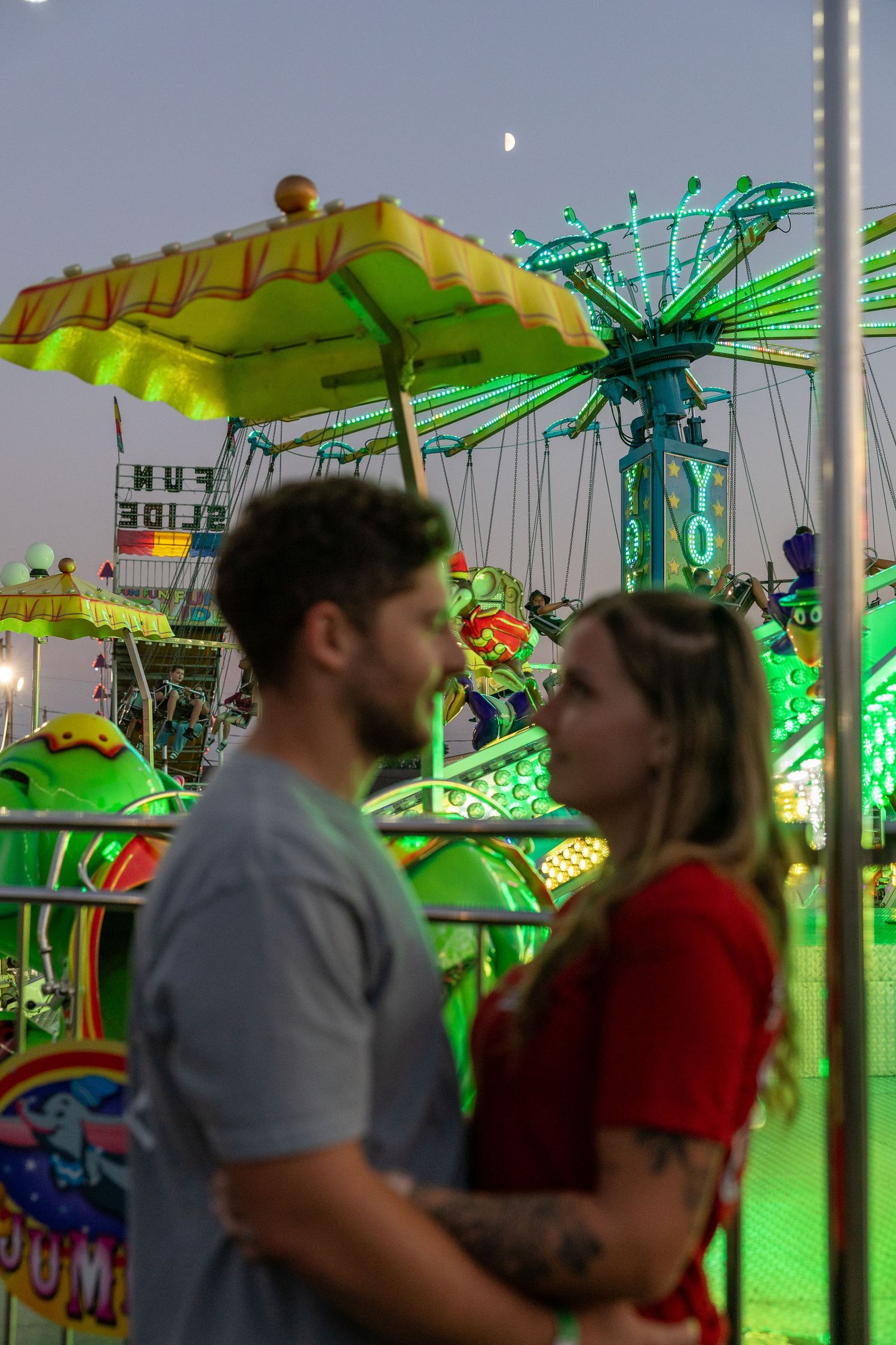 Couple standing close together at a colorful carnival fairground at dusk with glowing green rides in the background.