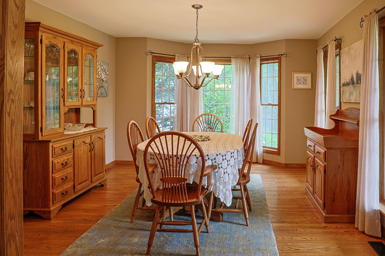 Elegant dining room with oak furniture, round table, Windsor chairs, hutch, hardwood floors, and bay windows with natural light.