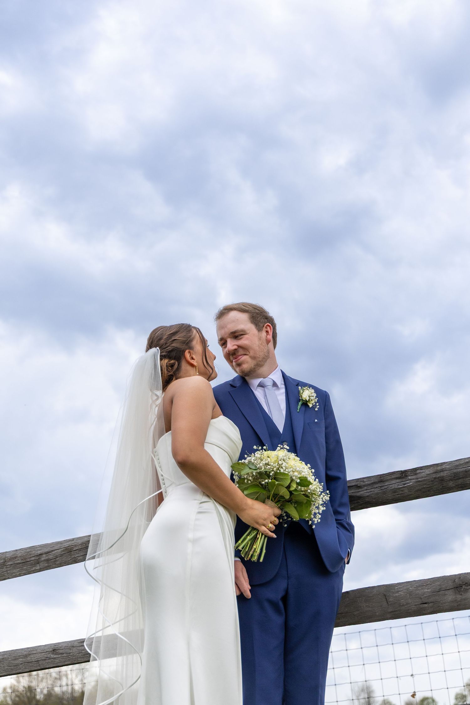 Bride and groom stand together outdoors by a wooden fence, she holds a white bouquet, both dressed in wedding attire under cloudy sky.