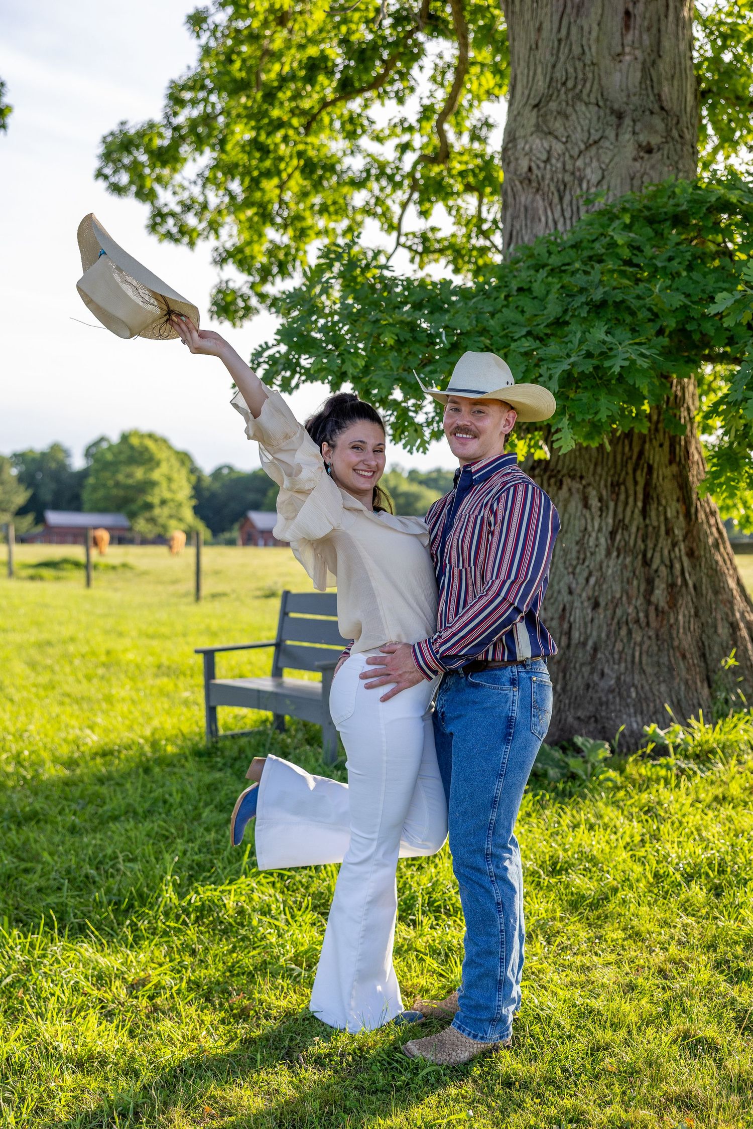 Joyful couple posing by a large tree, woman raising her hat while man holds her waist.