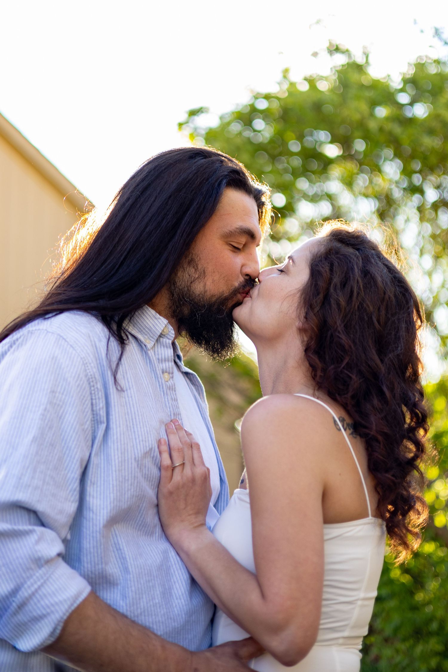 Bearded man with long hair kissing curly-haired woman outdoors.