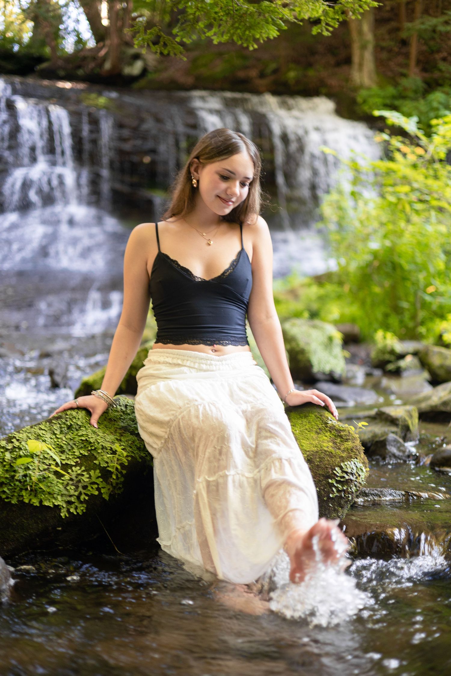 Young woman in black crop top and white skirt sitting on mossy rocks by a serene waterfall, splashing her feet in the stream.