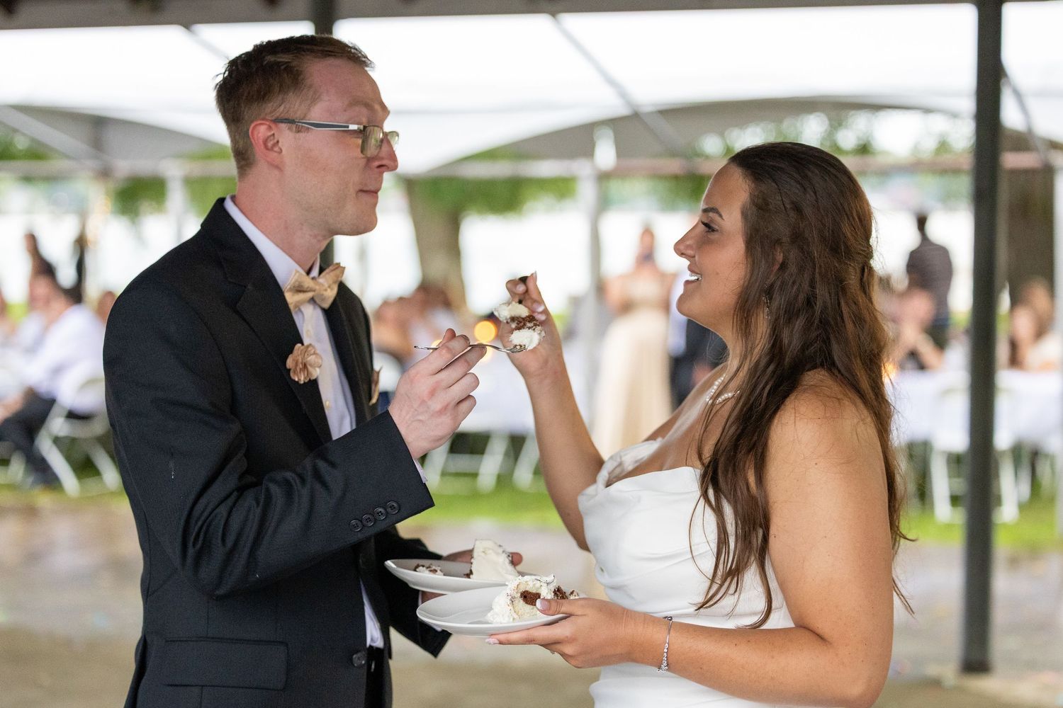 Bride and groom sharing wedding cake during outdoor reception ceremony, smiling joyfully at each other.