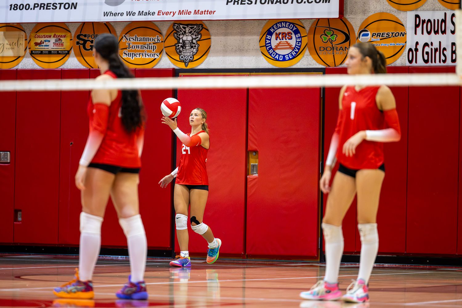High school girls volleyball players in red uniforms practice serving during an indoor gym match.